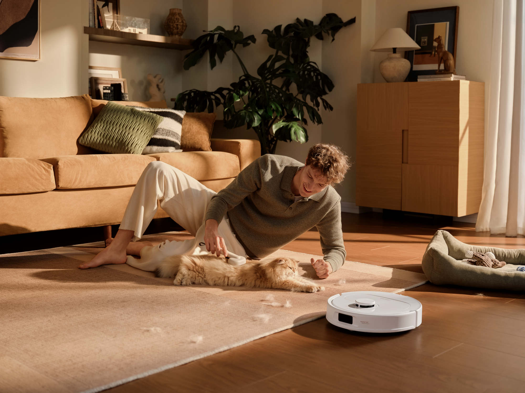 Egy férfi egy macskát kefél egy szőnyegen, miközben egy robotporszívó van a közelben.
