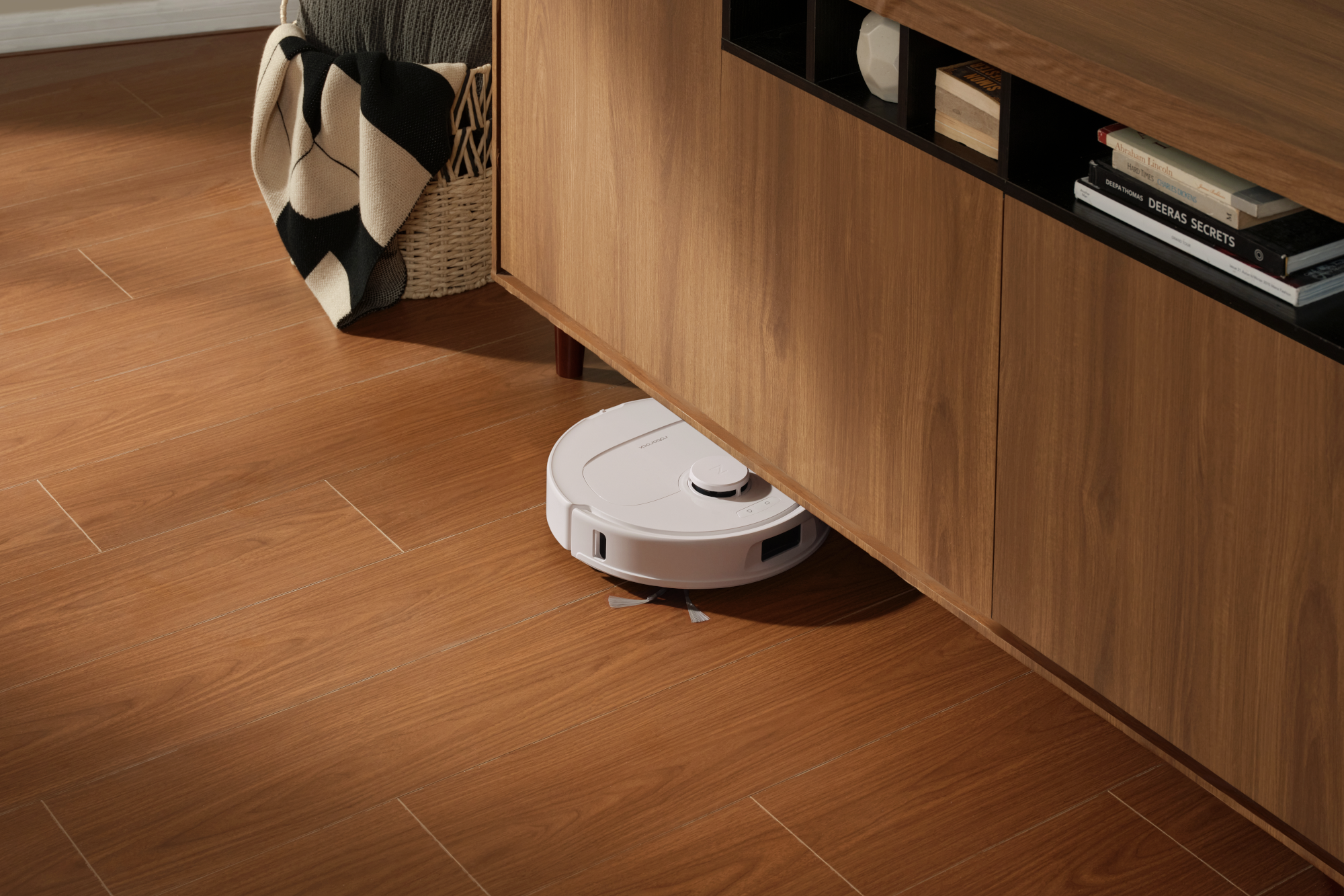 Egy fehér robotporszívó tisztítja a fapadlót egy fabútor alatt.