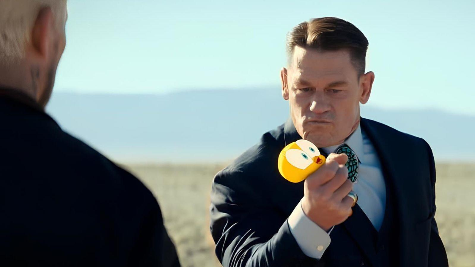 Ein Mann im Anzug in einer Wüste hält ein kleines gelbes Plüschtier mit großen blauen Augen.
