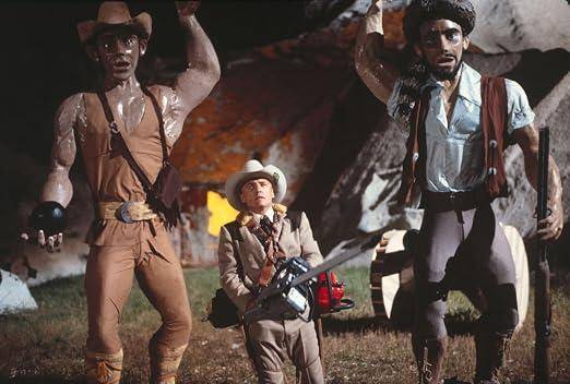 Ein kleiner Cowboy mit einer Kettensäge steht zwischen zwei großen Statuen vor einer Kulisse.