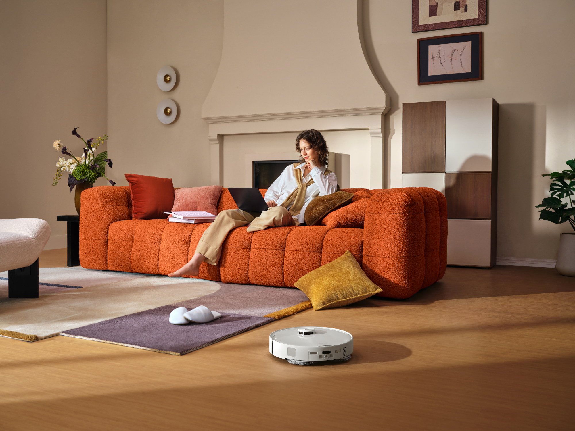 Egy nő narancssárga kanapén ül és laptopot használ, miközben egy robotporszívó gurul a padlón.