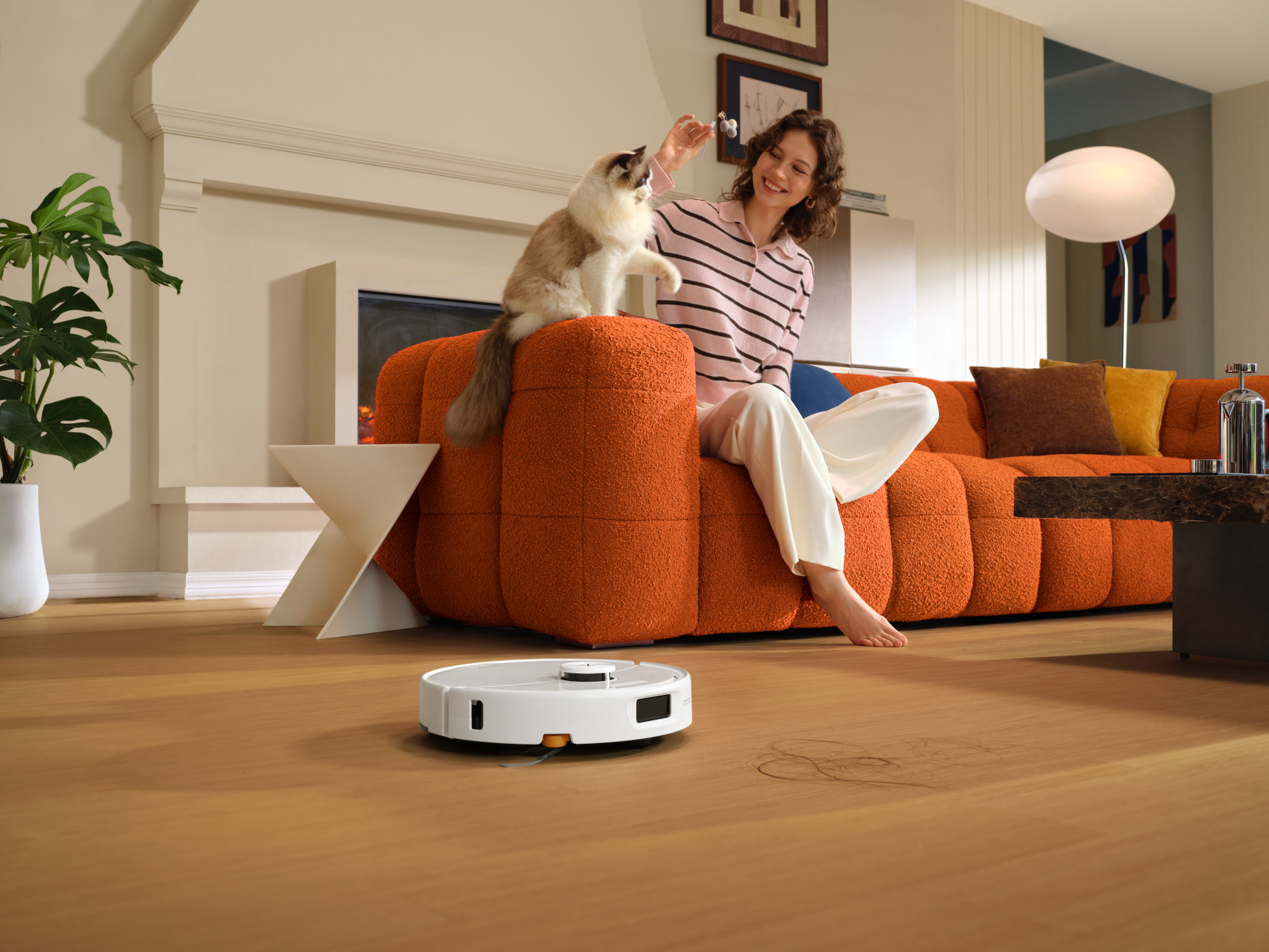 Egy nő narancssárga kanapén ül és a macskájával játszik, miközben egy robotporszívó tisztítja a fapadlót.