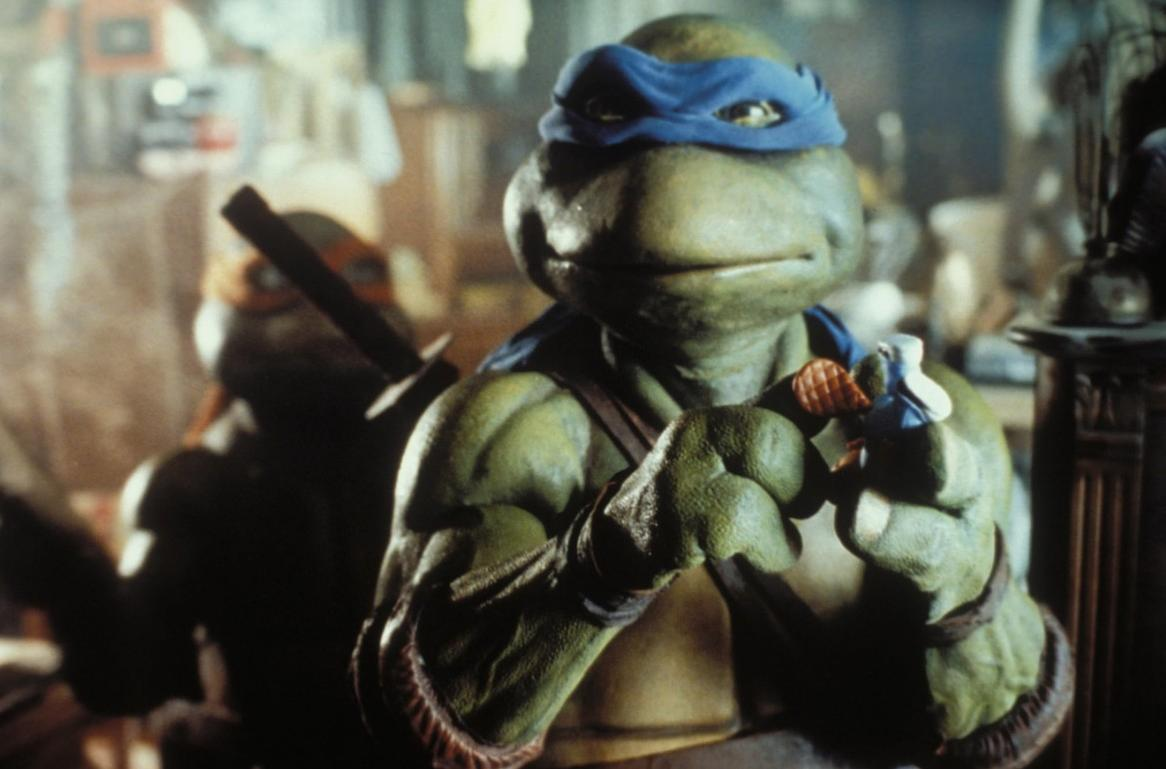 Eine Ninja-Schildkröte mit blauer Maske hält ein kleines Objekt, im Hintergrund steht eine weitere Schildkröte.