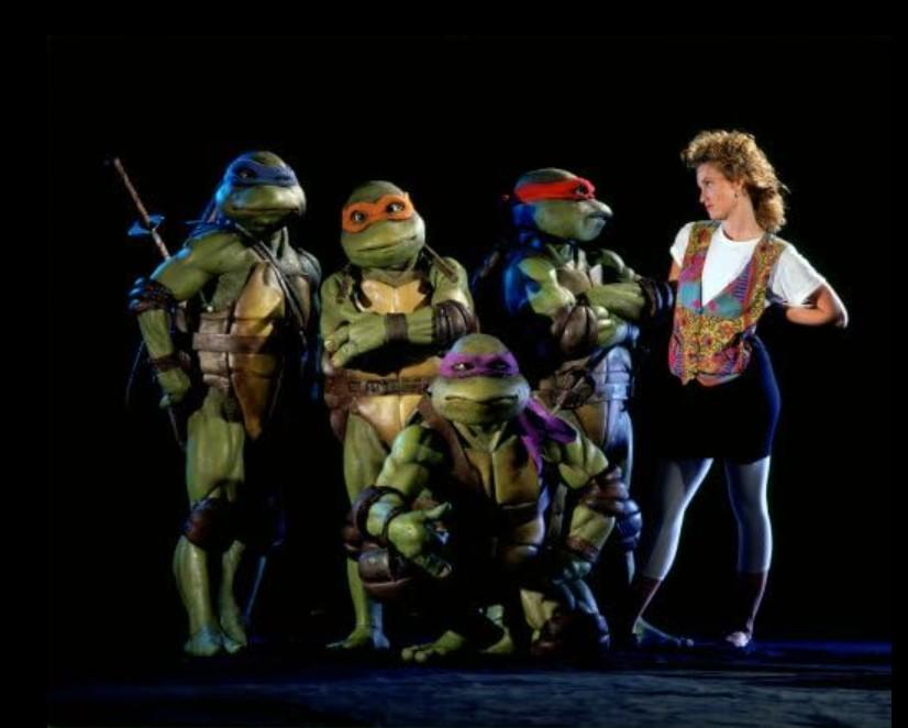 Vier Ninja Turtles in bunten Masken stehen zusammen mit einer Frau vor dunklem Hintergrund.