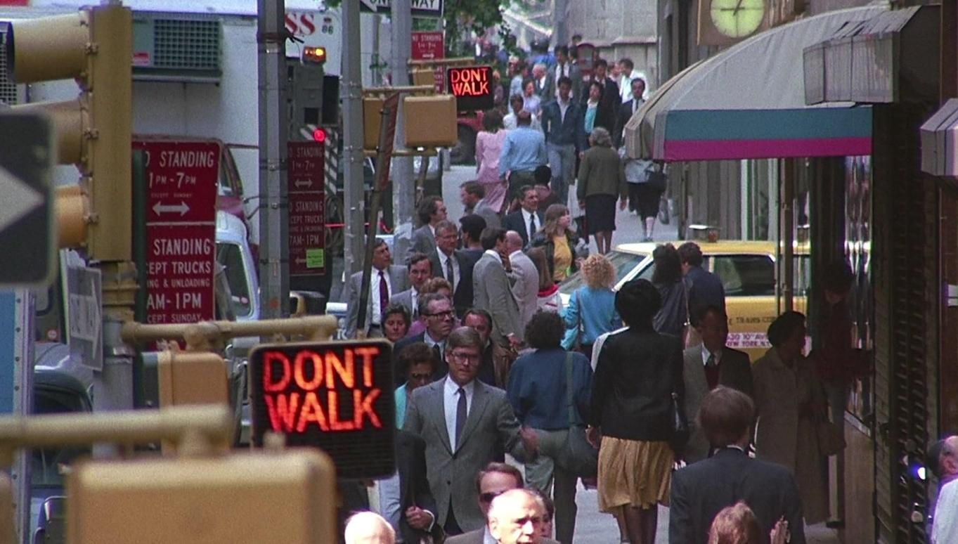 Menschenmenge geht an einem geschäftigen Stadtgehweg mit Straßenschildern und Geschäften vorbei.