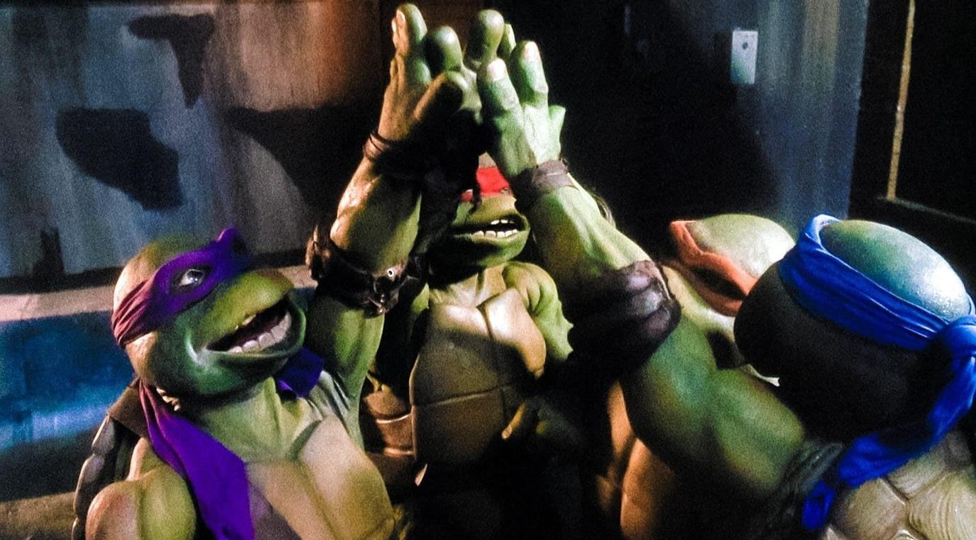 Vier Teenage Mutant Ninja Turtles geben sich im Dunkeln ein High-Five, während ihre bunten Masken im Scheinwerferlicht leuchten.
