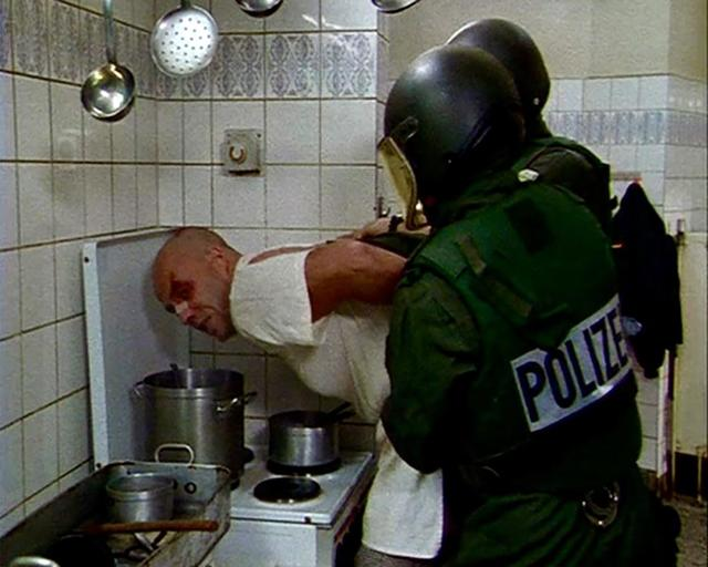 Zwei deutsche Polizeibeamte in Schutzkleidung fixieren einen Mann in einer gefliesten Küche.