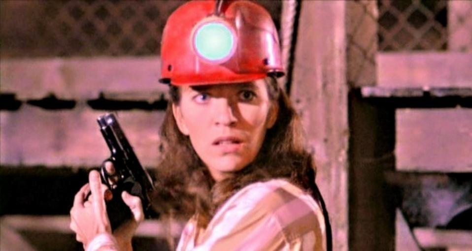 Eine Frau in Arbeitskleidung mit rotem Helm und leuchtender Lampe hält besorgt eine Waffe.
