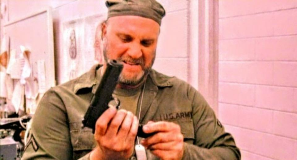 Ein Soldat in Uniform lächelt, während er in einem Raum mit rosafarbenen Wänden eine Waffe inspiziert.