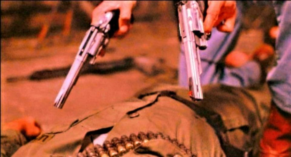 Zwei Hände halten Revolver, die auf eine am Boden liegende Person mit einem Bandolier gerichtet sind.