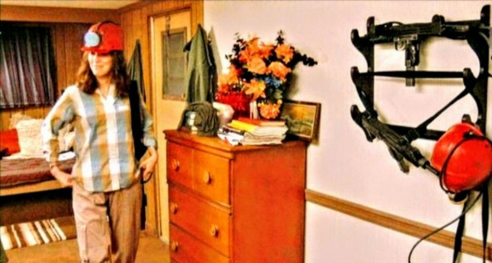 Eine Frau in kariertem Hemd und Schutzhelm steht an einer Kommode, ein Waffenhalter an der Wand.