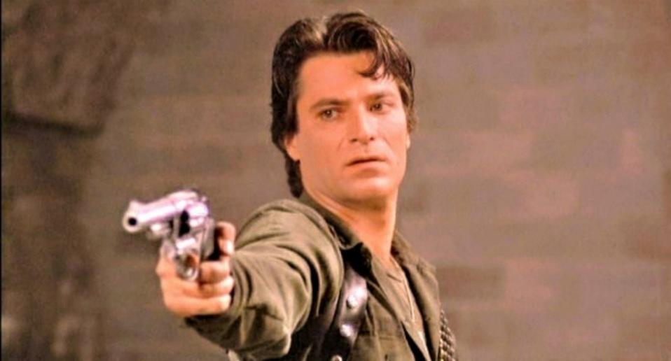 Ein Mann mit dunklem Haar zielt entschlossen mit einem silbernen Revolver in die Kamera.