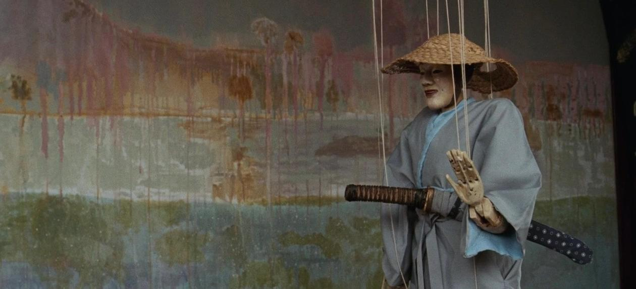 Eine traditionelle Marionette mit Strohhut, grauem Kimono und einem Schwert vor einem gemalten Hintergrund.