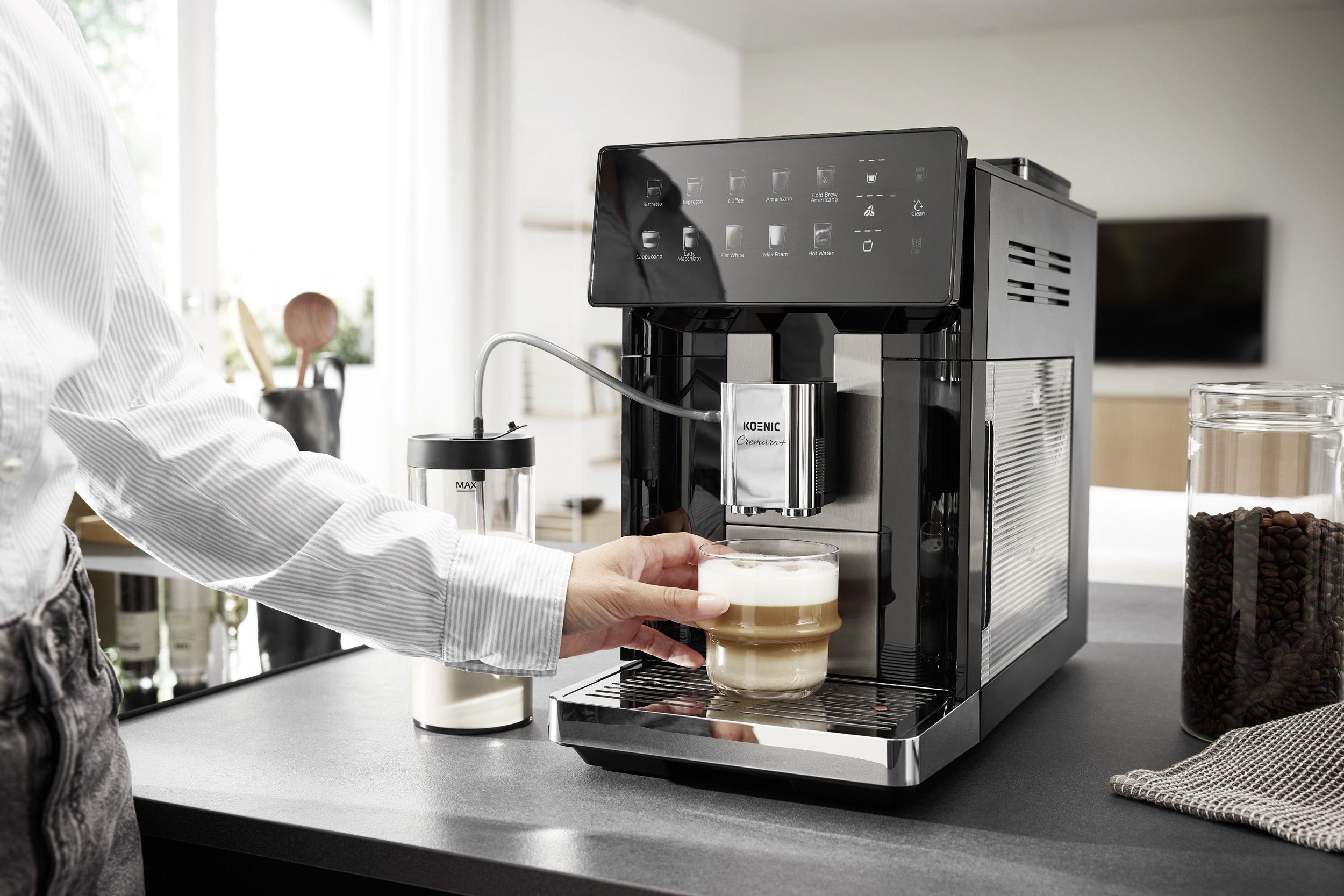 Eine Person stellt eine Tasse mit geschäumtem Kaffee unter den Auslauf eines modernen, schwarzen Kaffeeautomaten.