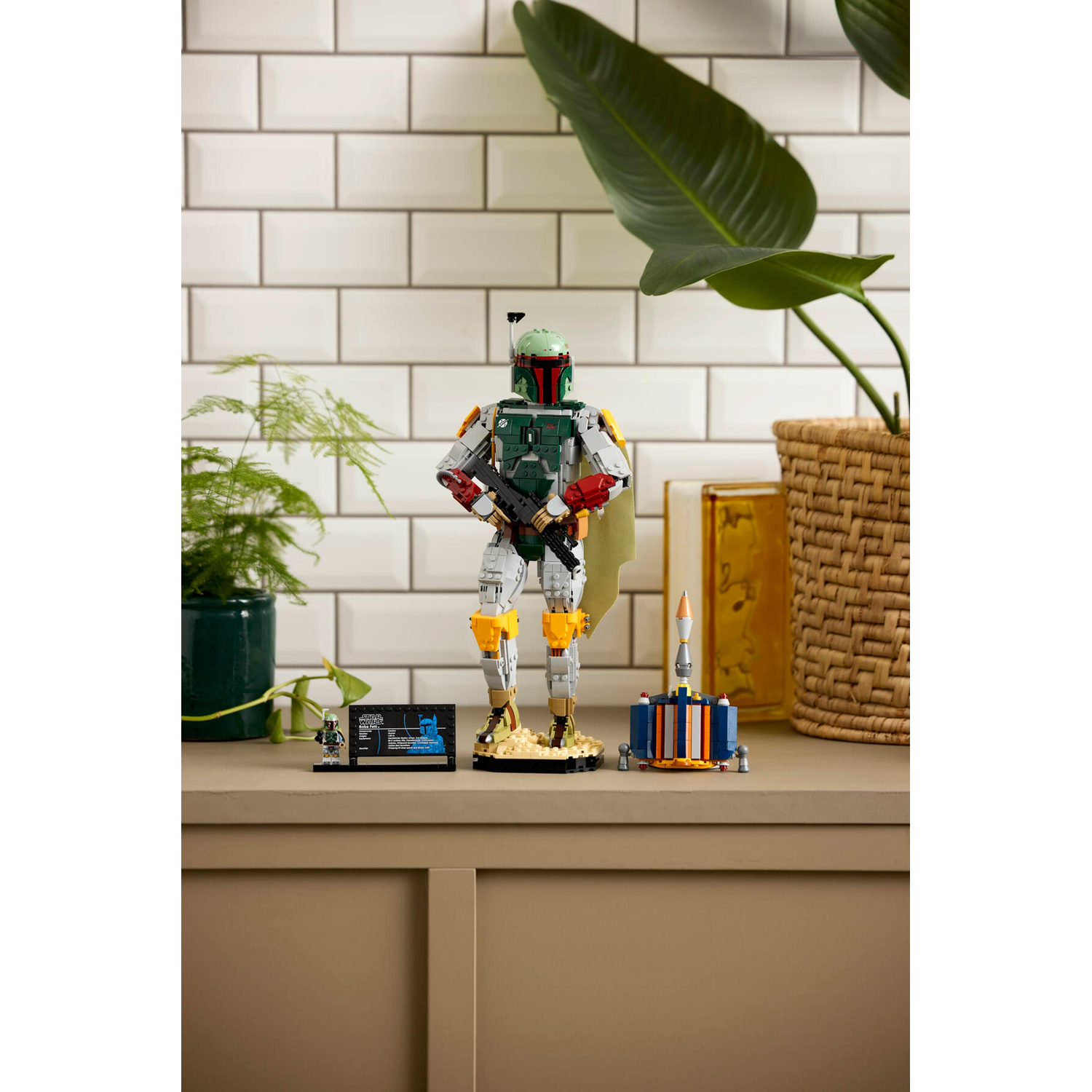 Una figura Lego di Boba Fett si trova su una mensola tra le piante, con un blaster, accanto a una piccola nave.