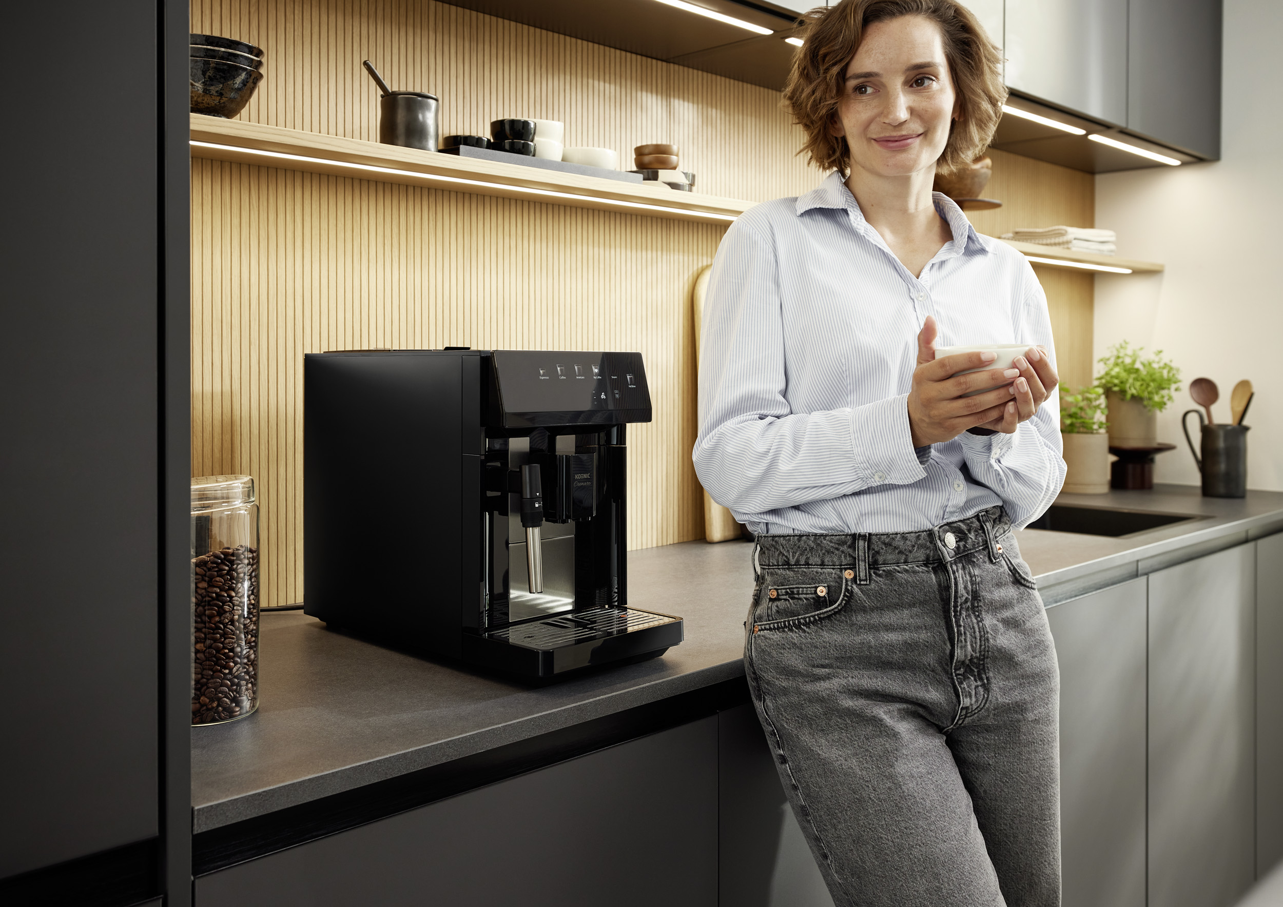Eine Frau hält eine Tasse in einer modernen Küche mit einer schwarzen Kaffeemaschine.
