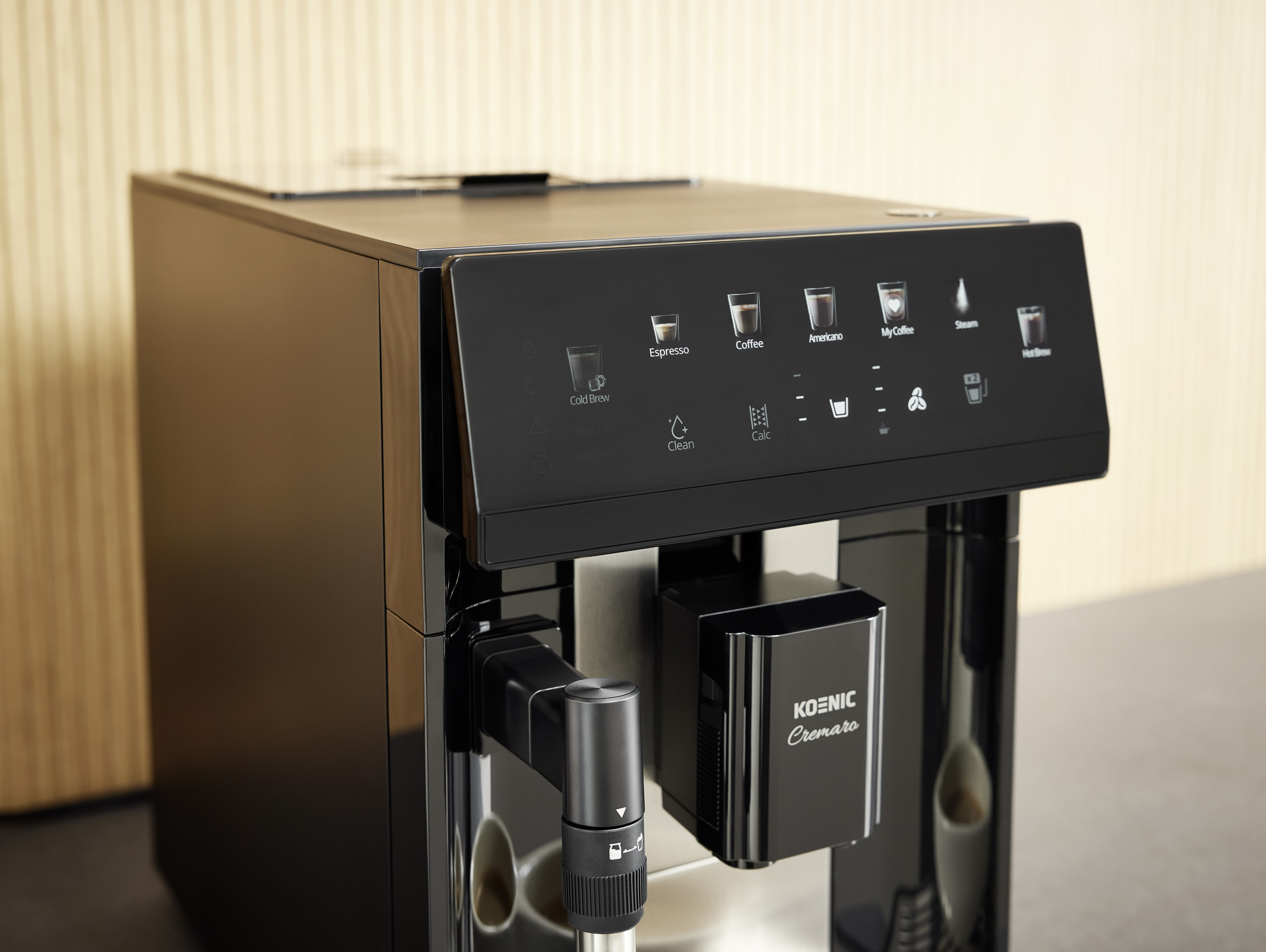 Schwarze Koenic Cremaro Kaffeevollautomat mit Touch-Bedienfeld und Milchaufschäumer vor Holzwand.