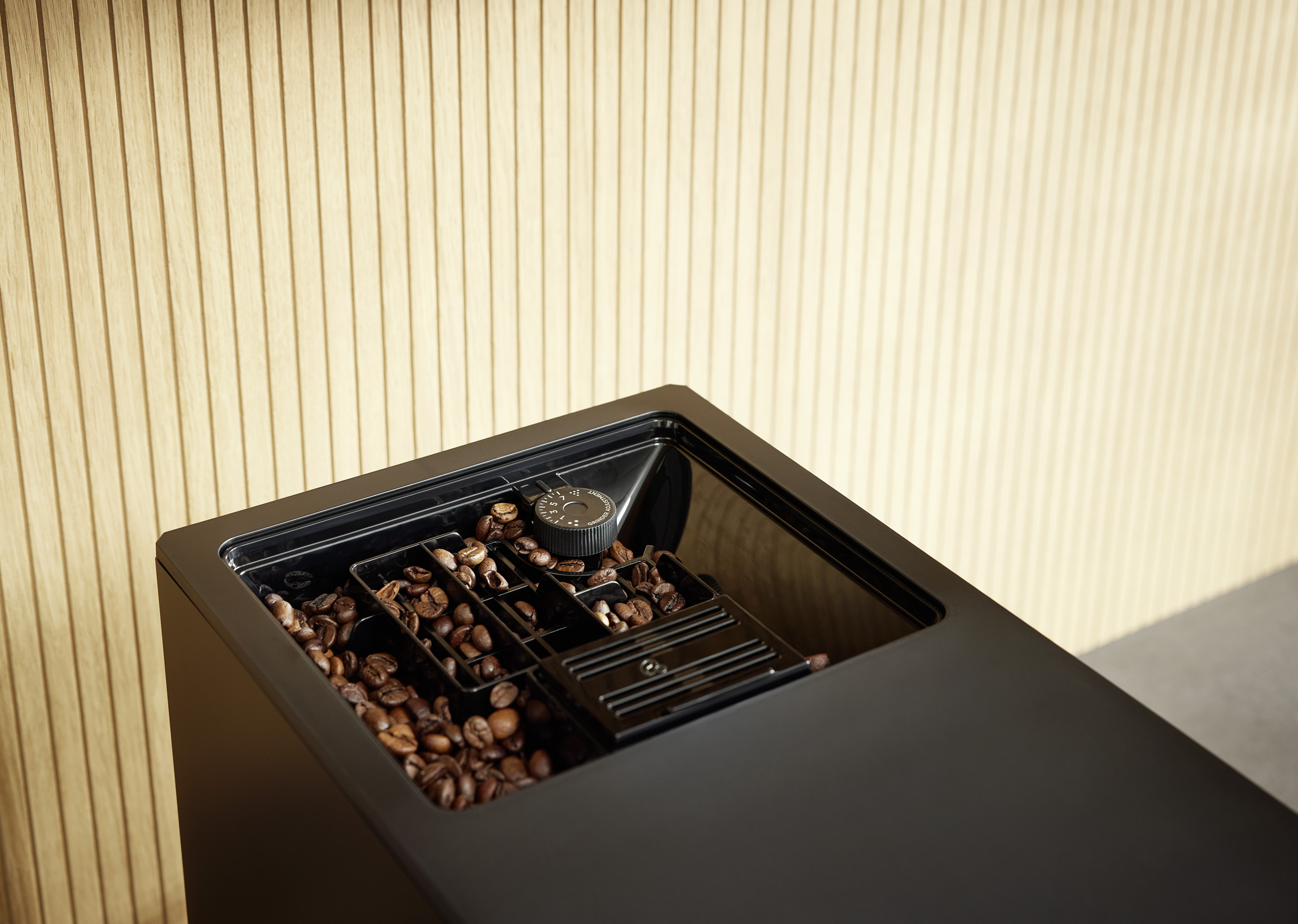 Kaffeebohnen im Mahlwerk einer modernen schwarzen Kaffeemaschine vor Holzwand.