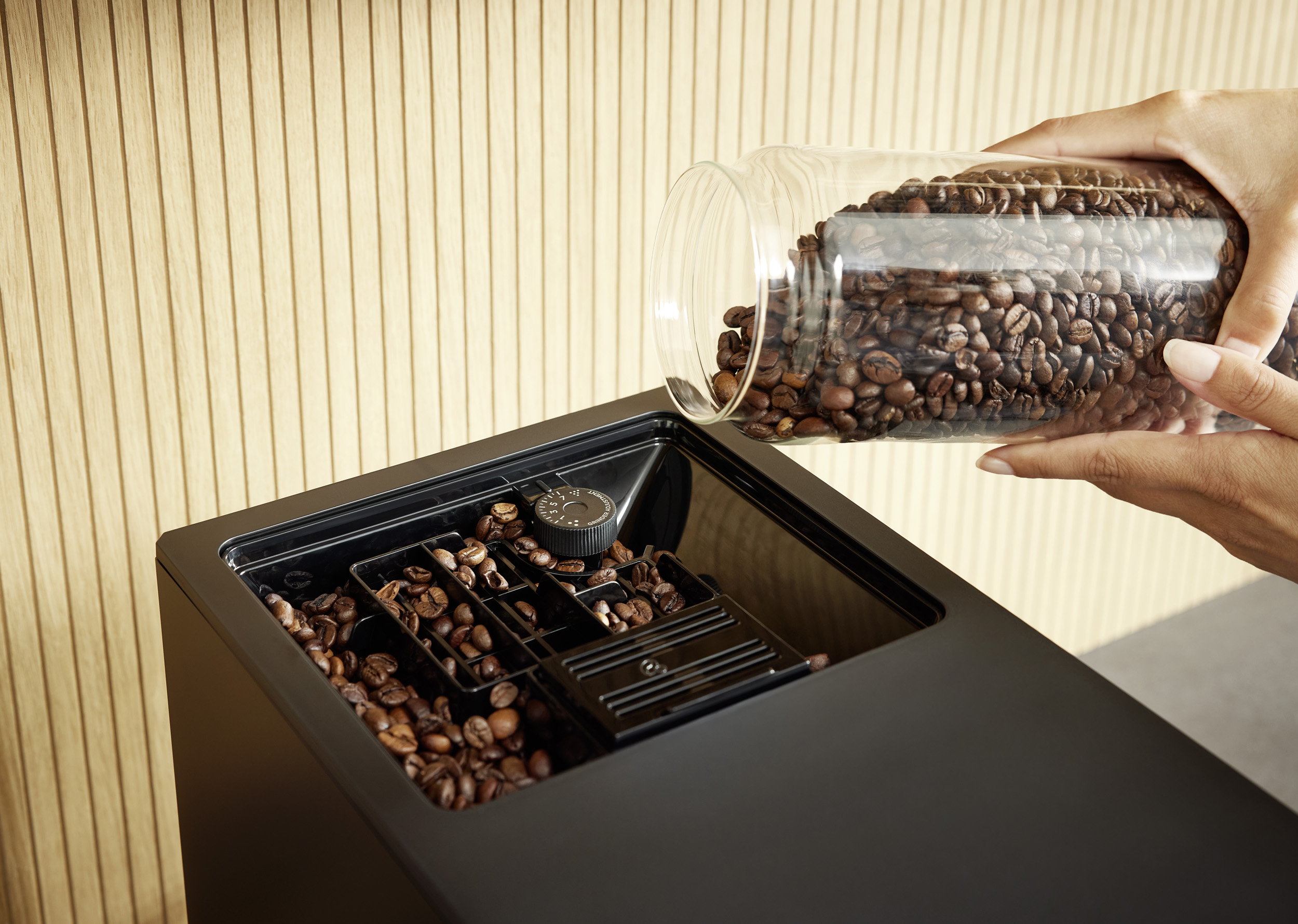 Jemand schüttet Kaffeebohnen aus einem Glasbehälter in das Mahlwerk einer Kaffeemaschine.