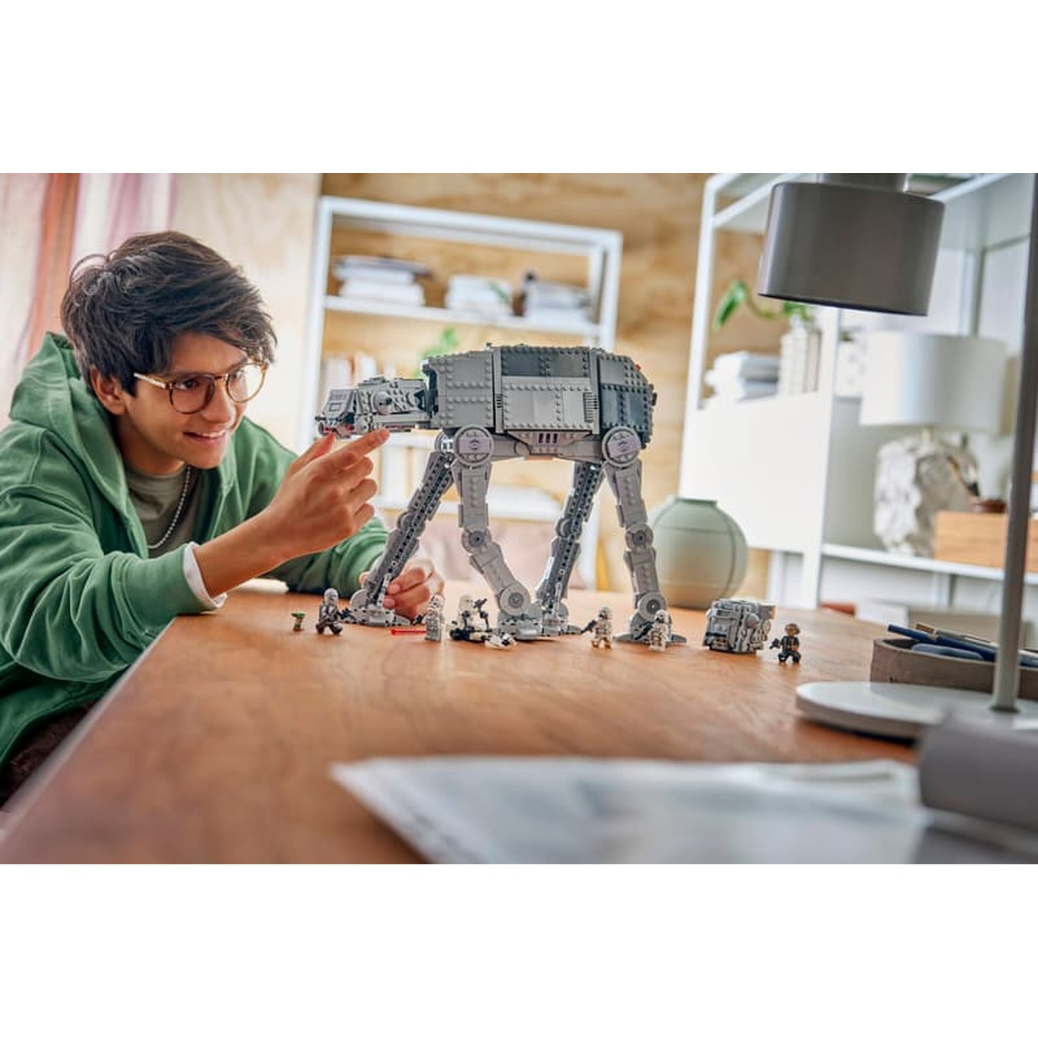 Un ragazzo con gli occhiali e felpa verde gioca con un camminatore Lego AT-ST grigio su un tavolo di legno.