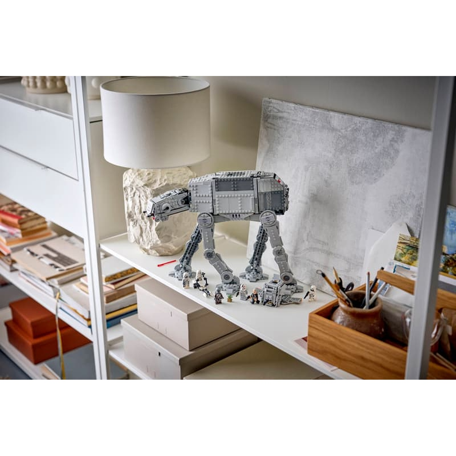 Un modello Lego AT-AT Walker su uno scaffale vicino a una lampada, un dipinto e matite in un supporto di legno.