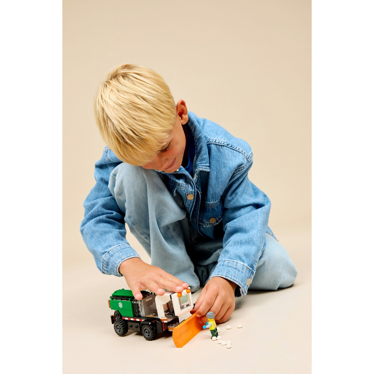 Un bambino biondo in jeans gioca con un veicolo Lego e una piccola statuetta su sfondo chiaro.