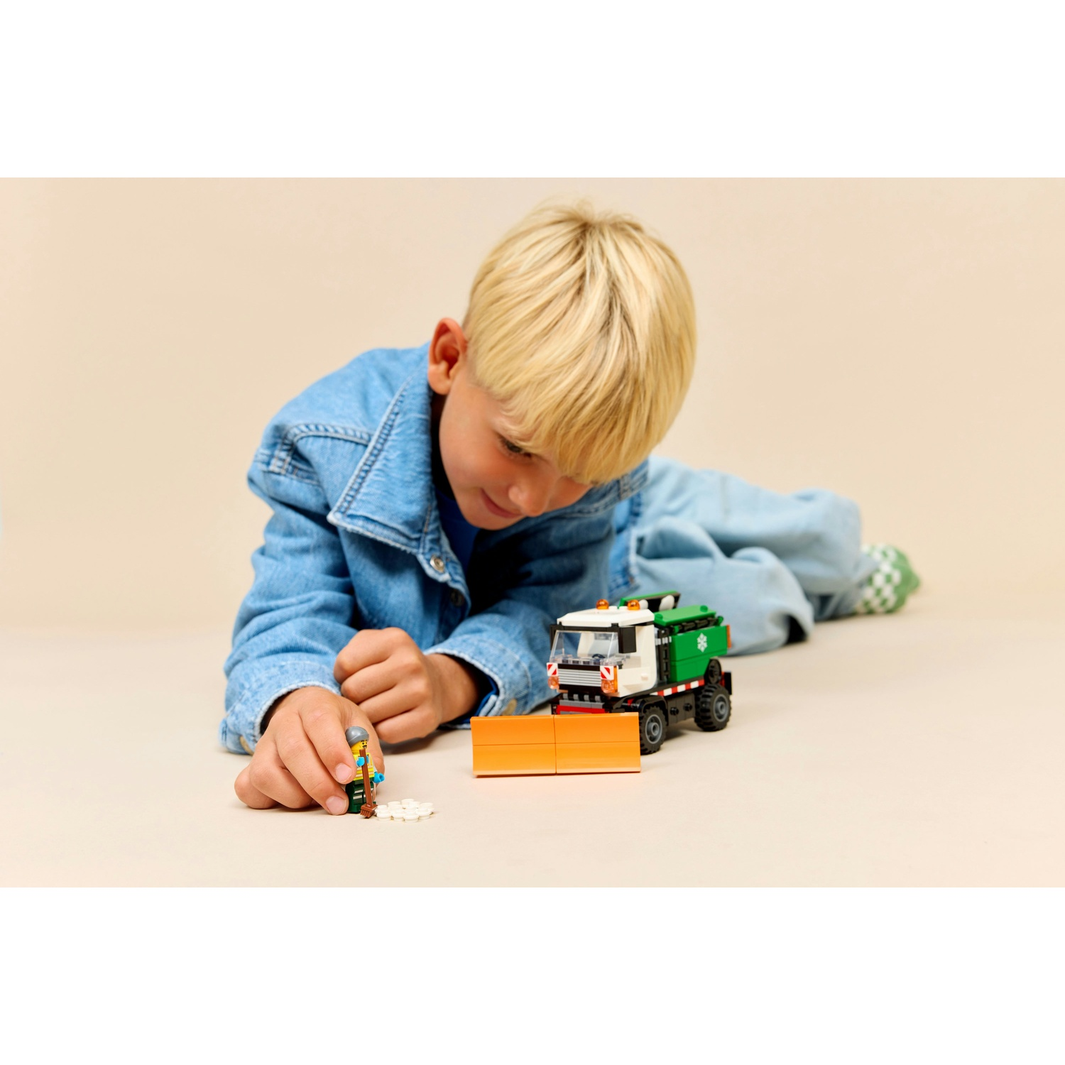 Un bambino biondo con giacca di jeans gioca a terra con uno spazzaneve Lego e una minifigura.