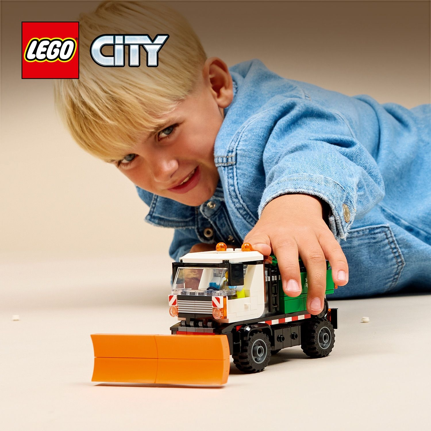 Un bambino biondo gioca con un camion giocattolo spazzaneve Lego City in primo piano.