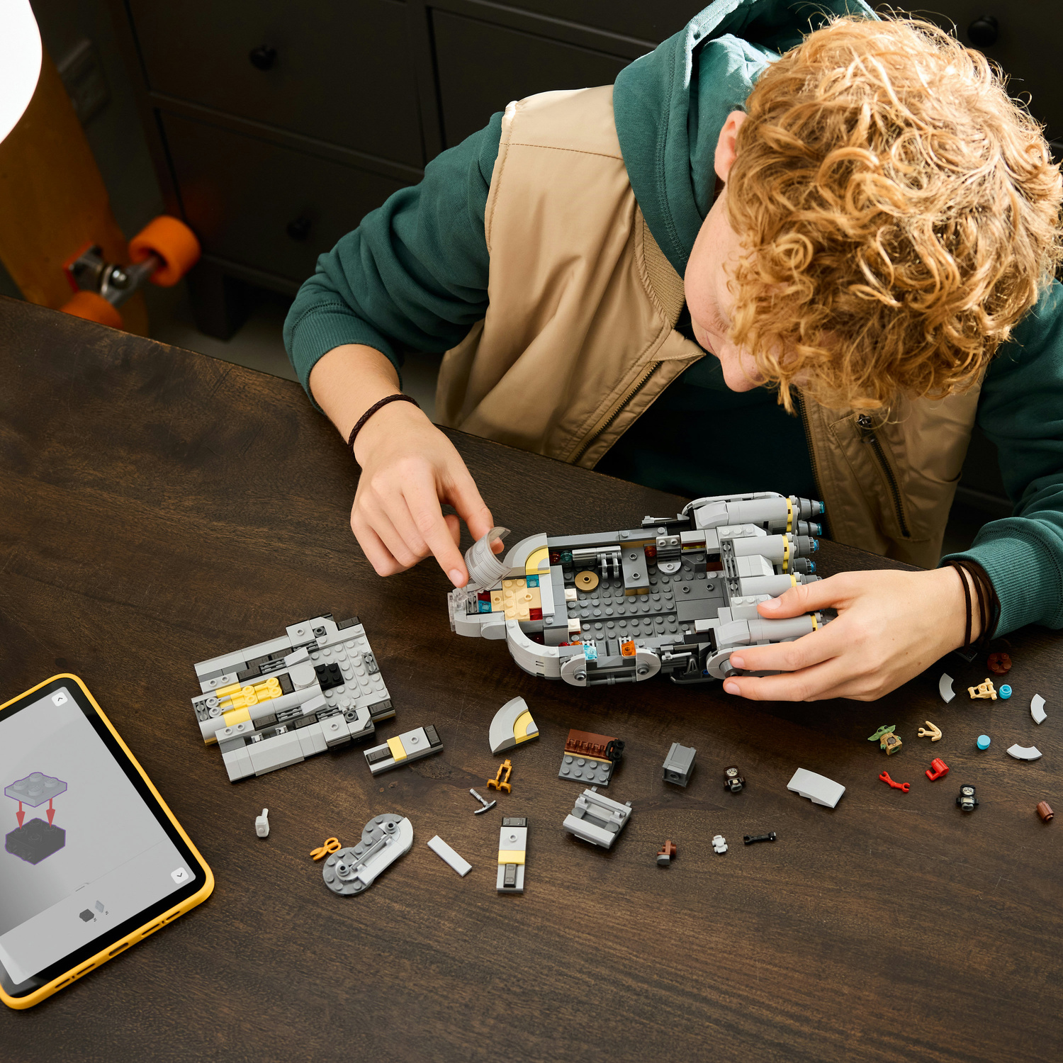 Un ragazzo riccio costruisce un'astronave Lego grigia su un tavolo di legno scuro con istruzioni digitali su un tablet.