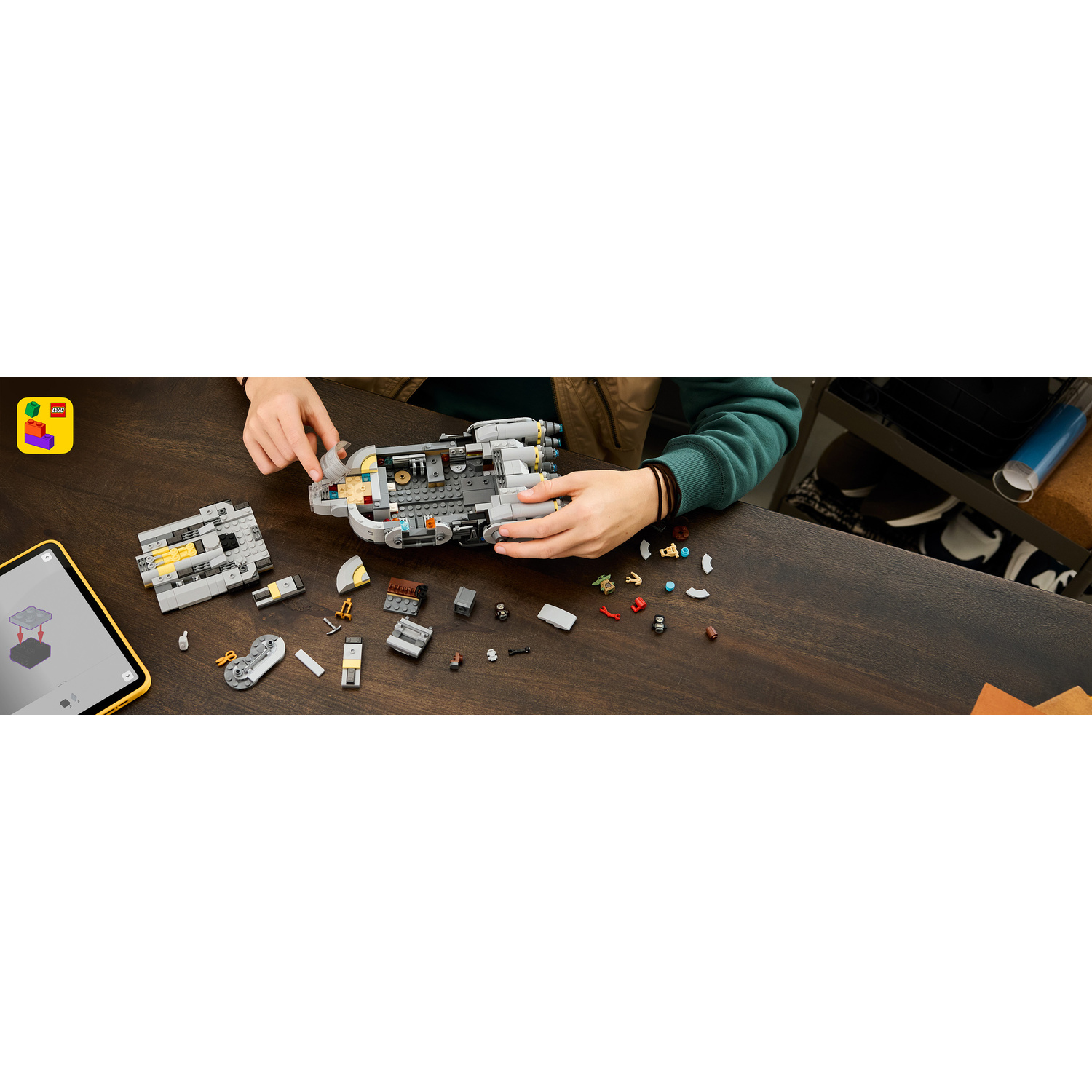 Qualcuno costruisce un modello Lego grigio su una scrivania di legno, con un tablet e pezzi.