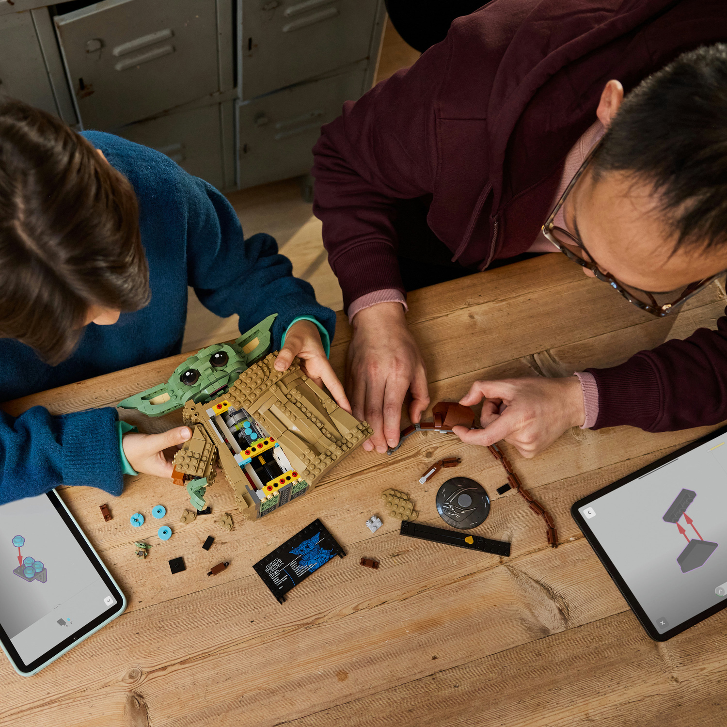Un adulto e un bambino costruiscono insieme un modello Lego di Grogu su un tavolo di legno, guidati da tablet.