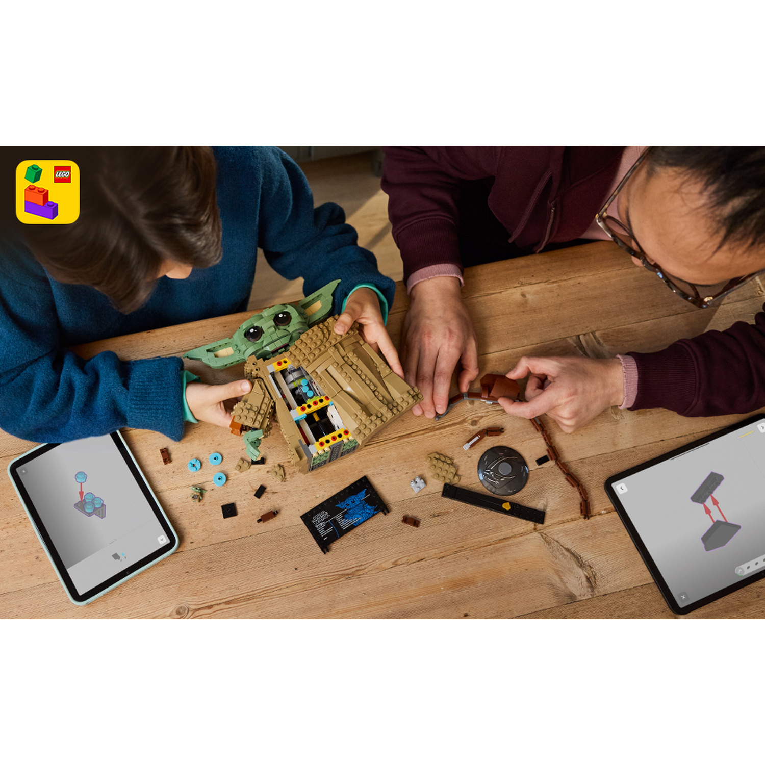 Un bambino e un adulto costruiscono insieme una figura Lego Star Wars Grogu su un tavolo di legno con tablet.