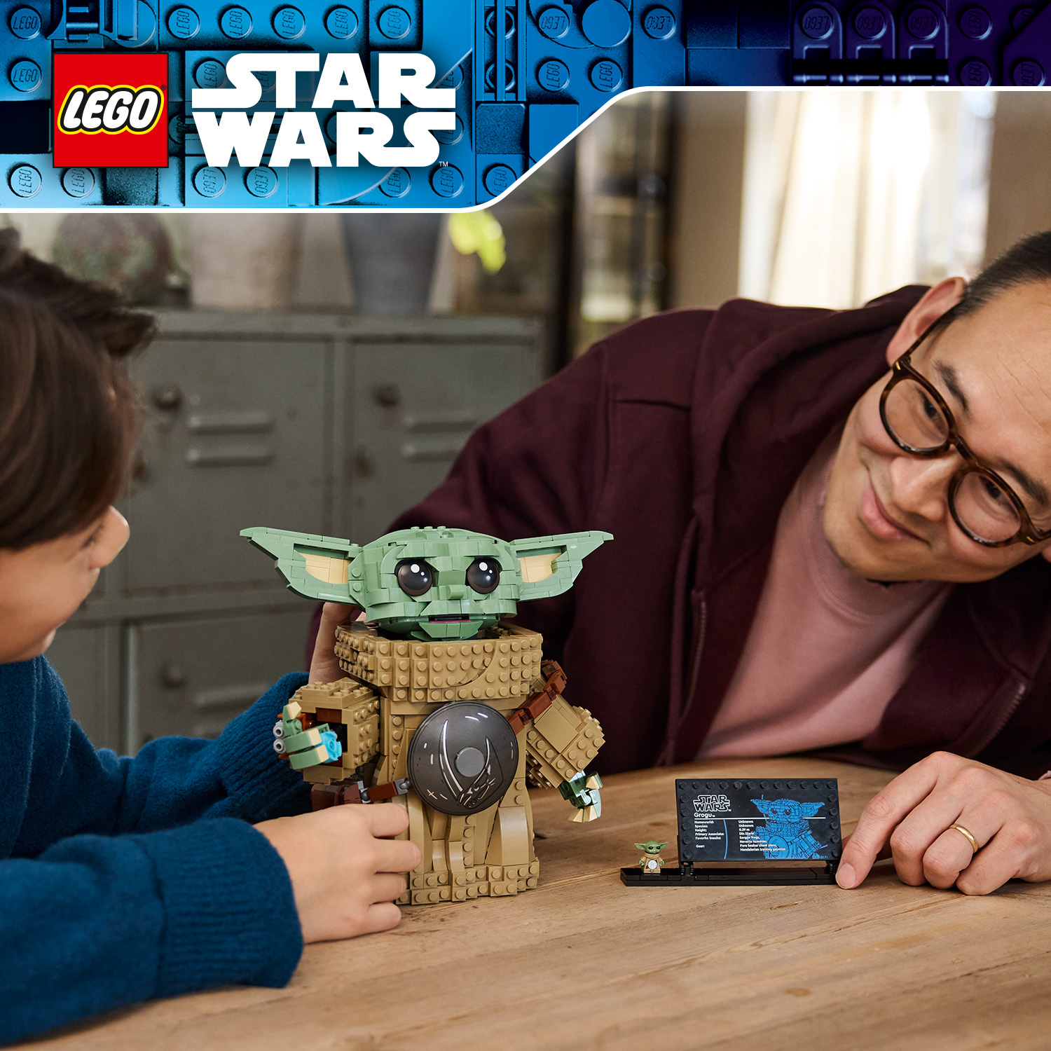 Un bambino e un adulto assemblano insieme una statuetta Lego Star Wars Grogu su un tavolo di legno.
