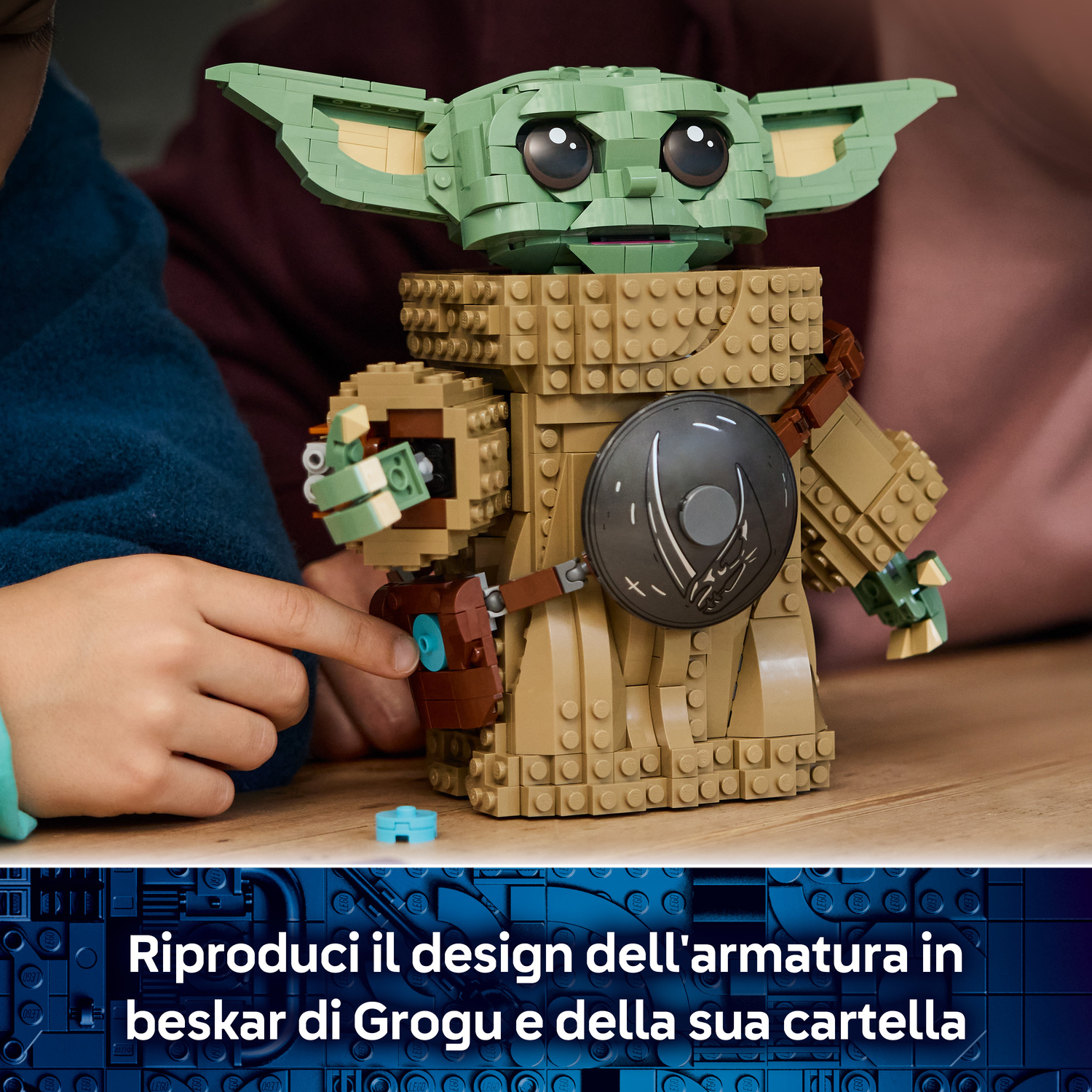 Un bambino tocca una statuetta Lego di Grogu di Star Wars, con cappotto marrone e scudo rotondo.
