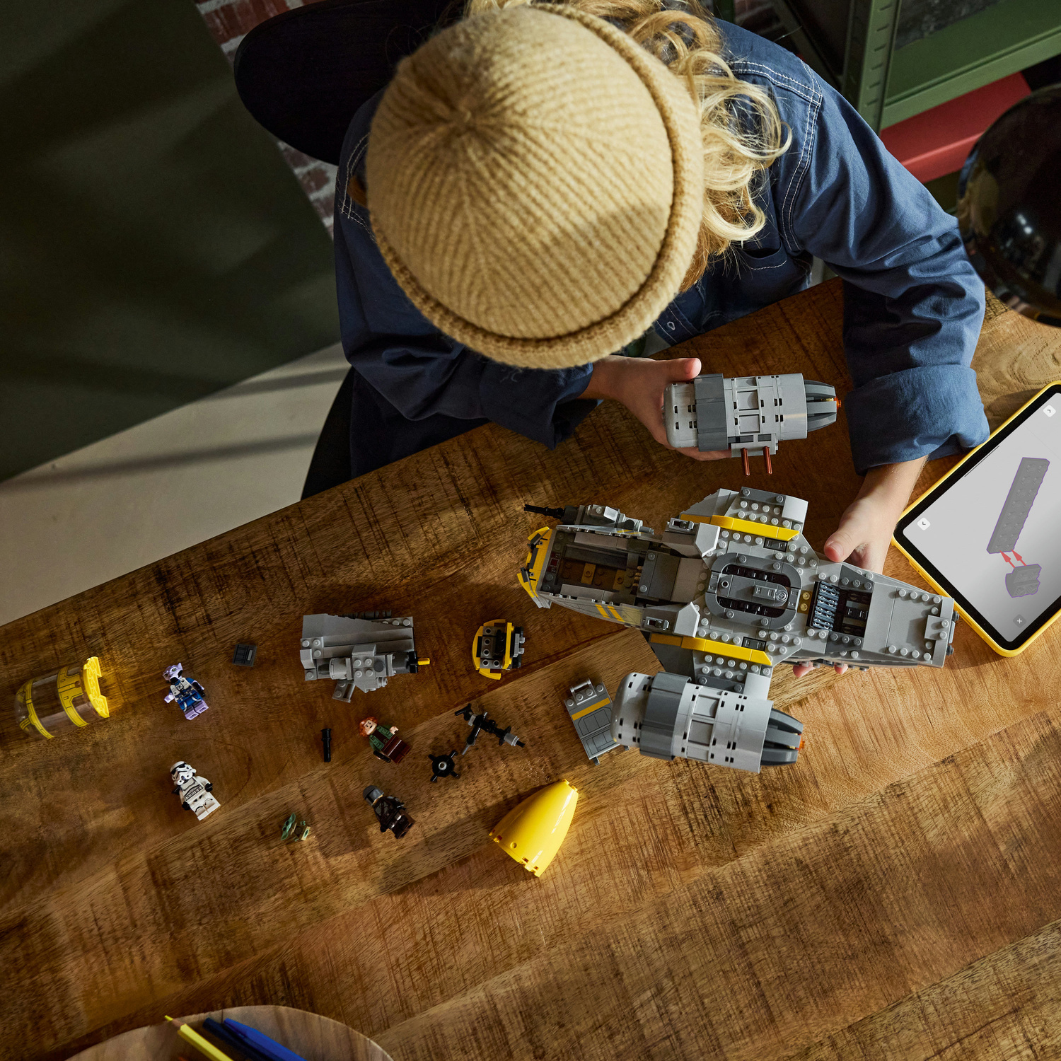 Una persona con berretto giallo costruisce un'astronave Lego grigia su un tavolo di legno con minifigure e tablet.