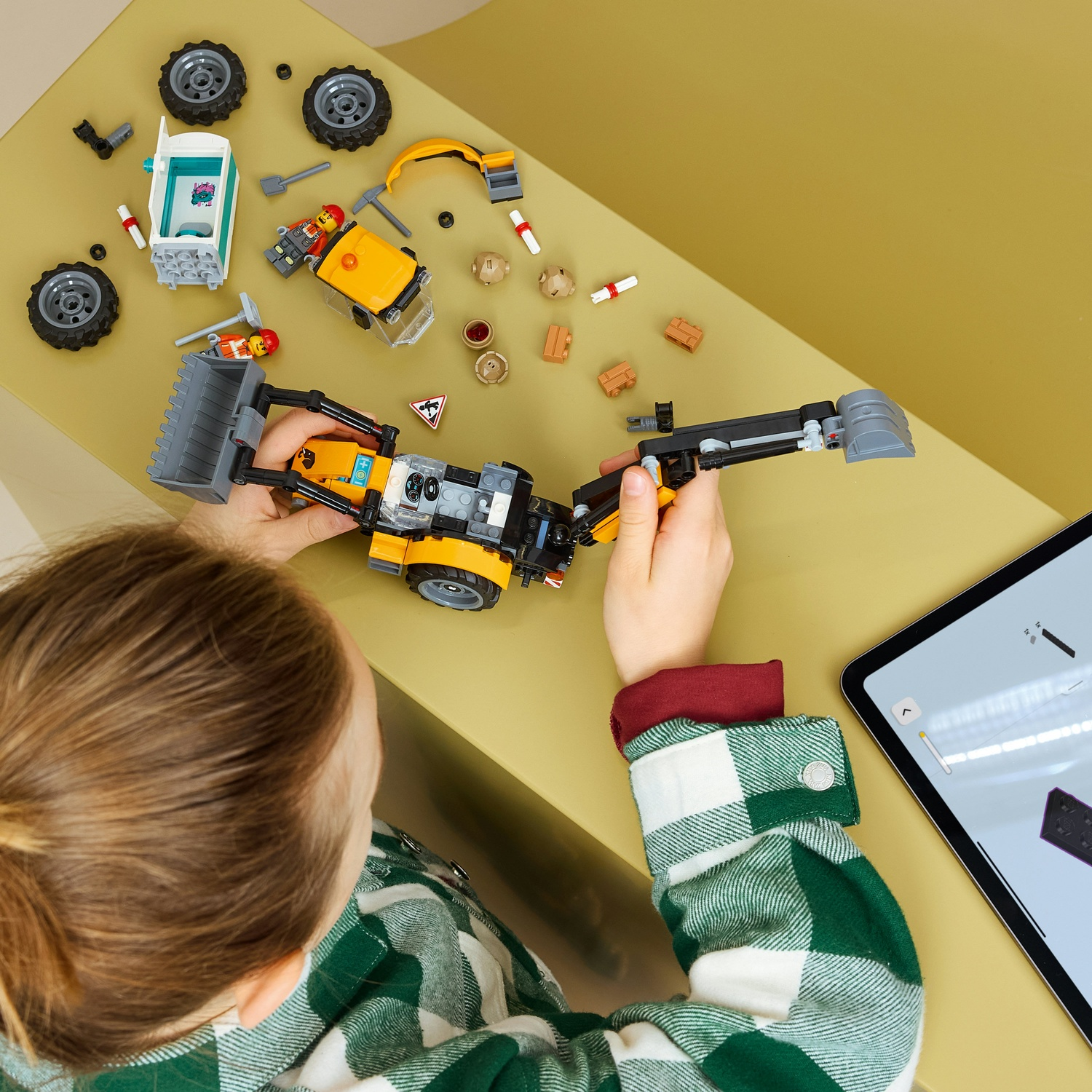 Un bambino con una camicia a quadri verde costruisce un trattore Lego giallo seguendo le istruzioni su un tablet.