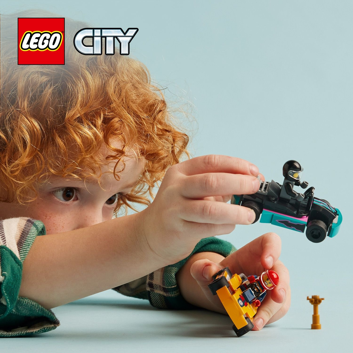 Un bambino con i capelli rossi ricci gioca con i kart LEGO City su sfondo azzurro.
