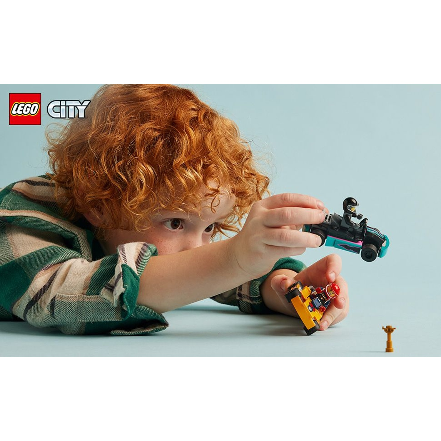 Un bambino dai capelli rossi gioca con macchinine Lego City su sfondo azzurro.