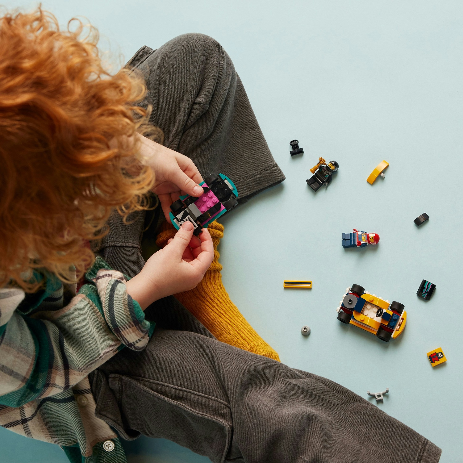 Un bambino con i capelli rossi siede sul pavimento giocando con mattoncini Lego colorati e macchinine giocattolo.