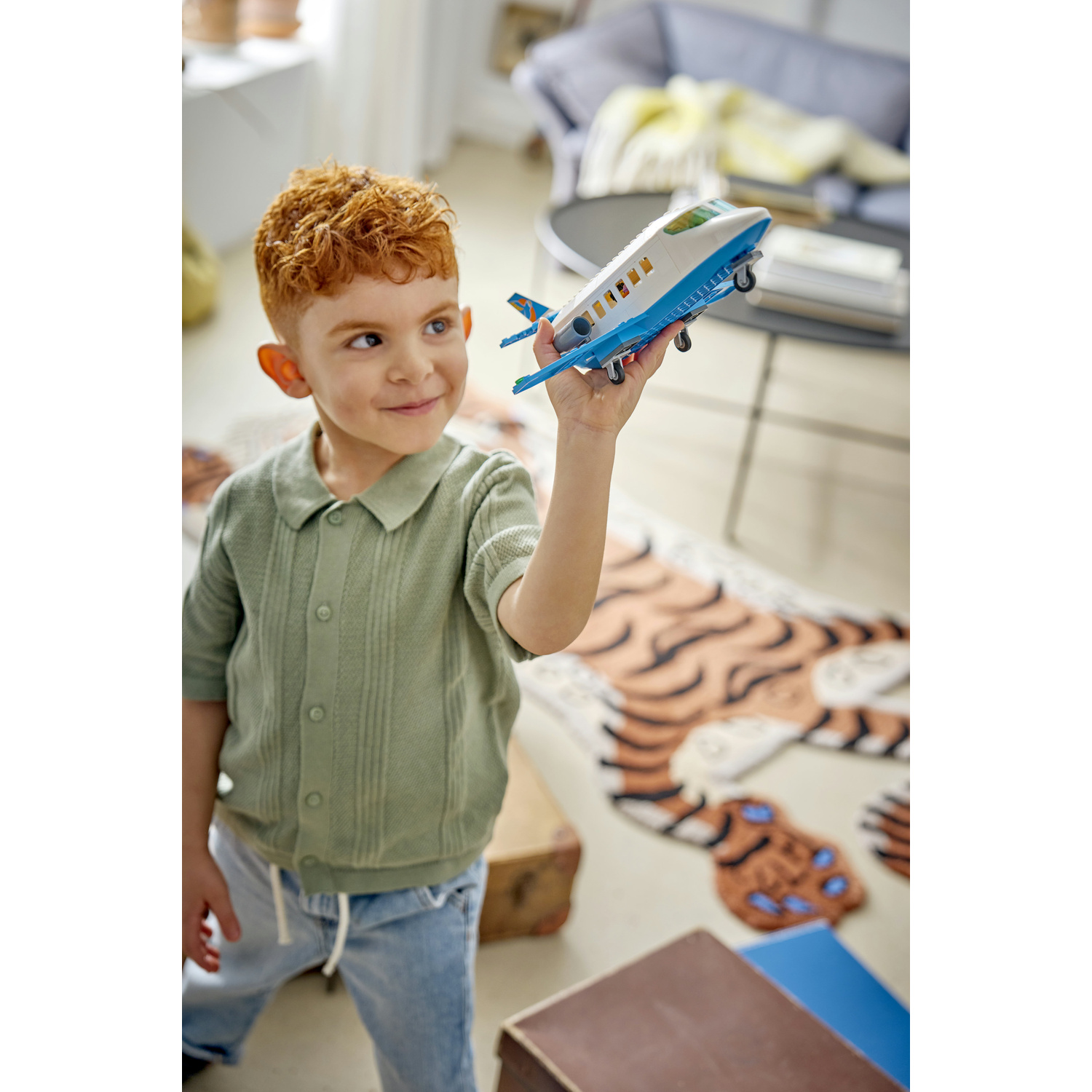 Un bambino dai capelli rossi con camicia verde gioca con un aereo giocattolo blu in una stanza.