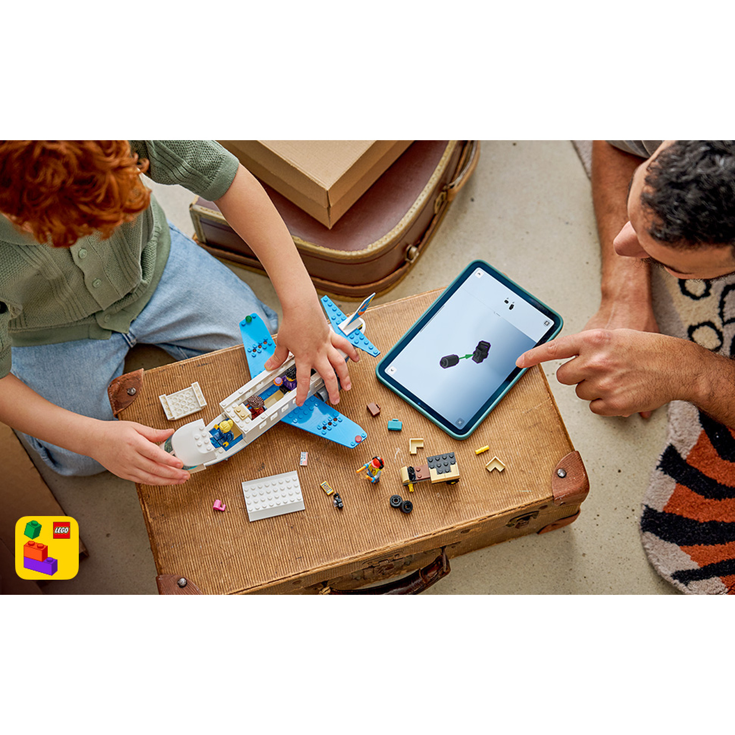 Un bambino e un adulto costruiscono un aereo Lego blu e bianco guardando un tablet.