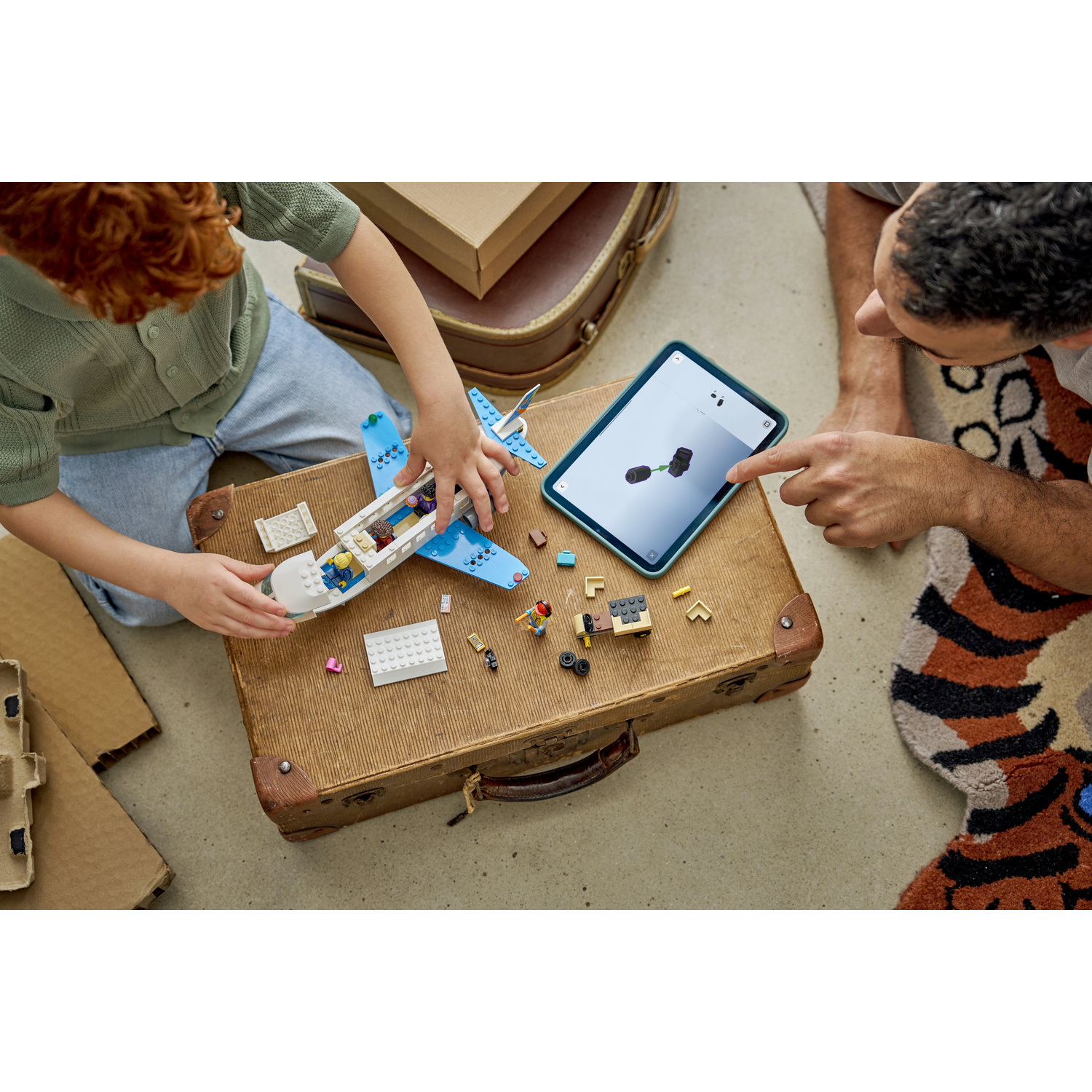 Un bambino e un adulto costruiscono un aereo Lego su una vecchia valigia, mentre un tablet mostra le istruzioni.