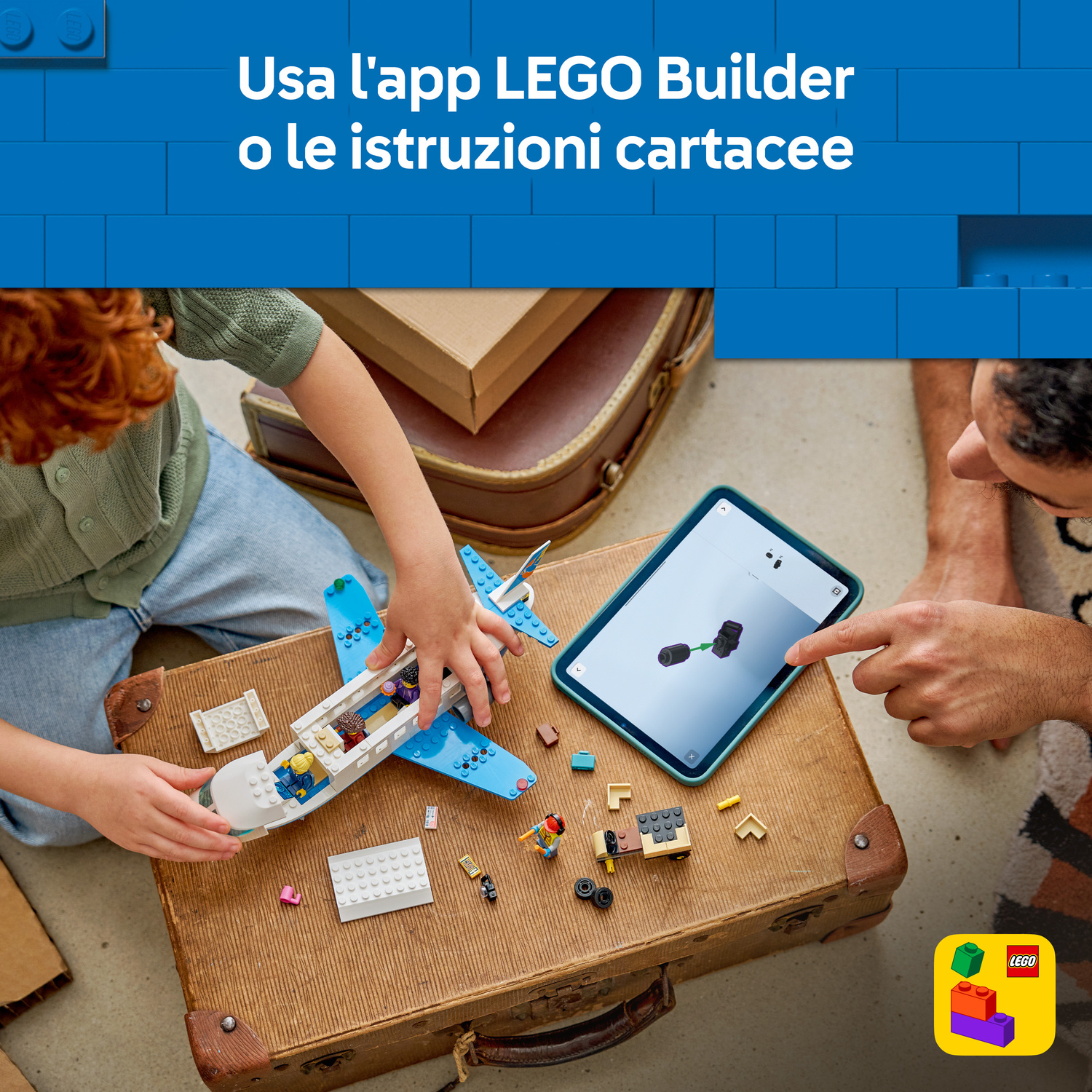 Bambino costruisce aereo Lego su valigia mentre adulto indica tablet.