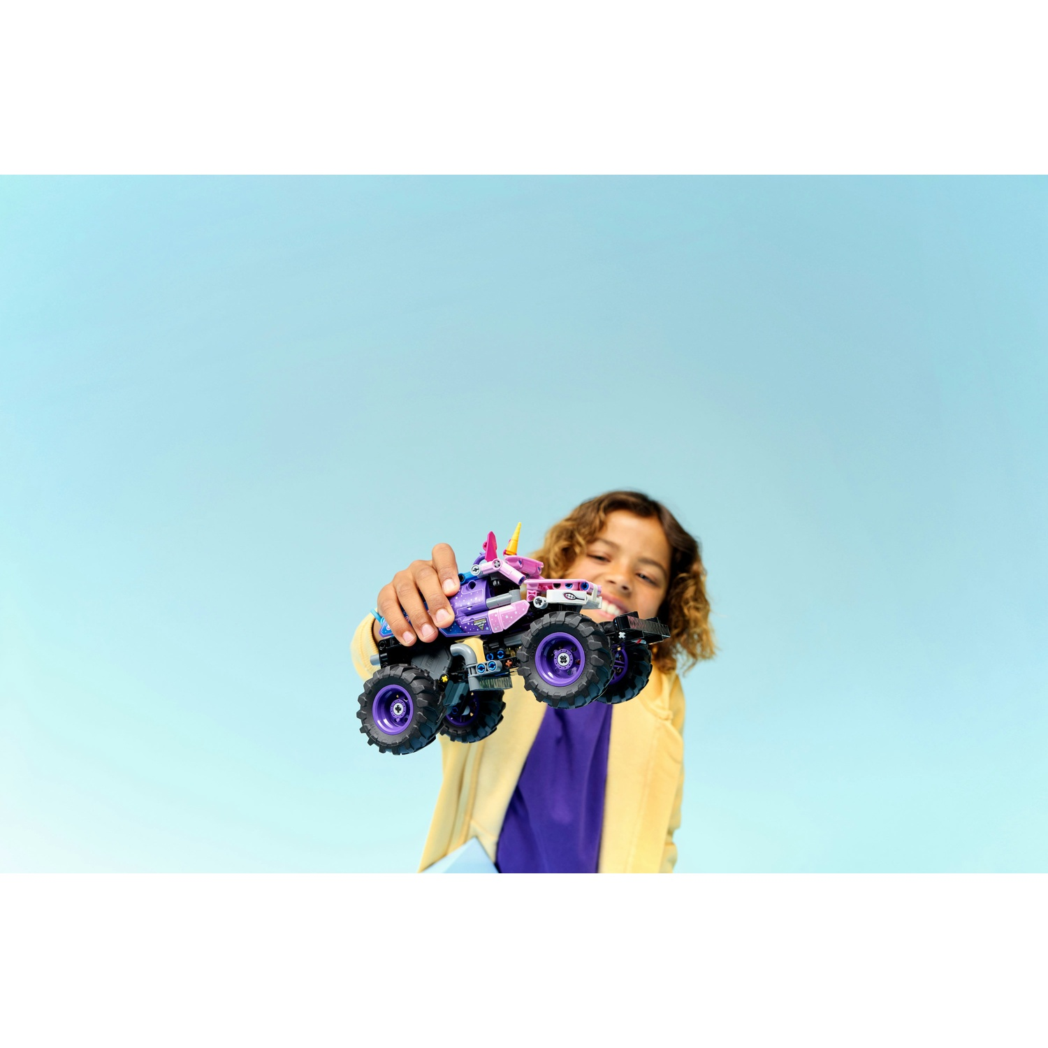 Un bambino sorridente tiene in mano un monster truck giocattolo viola e rosa su sfondo azzurro.