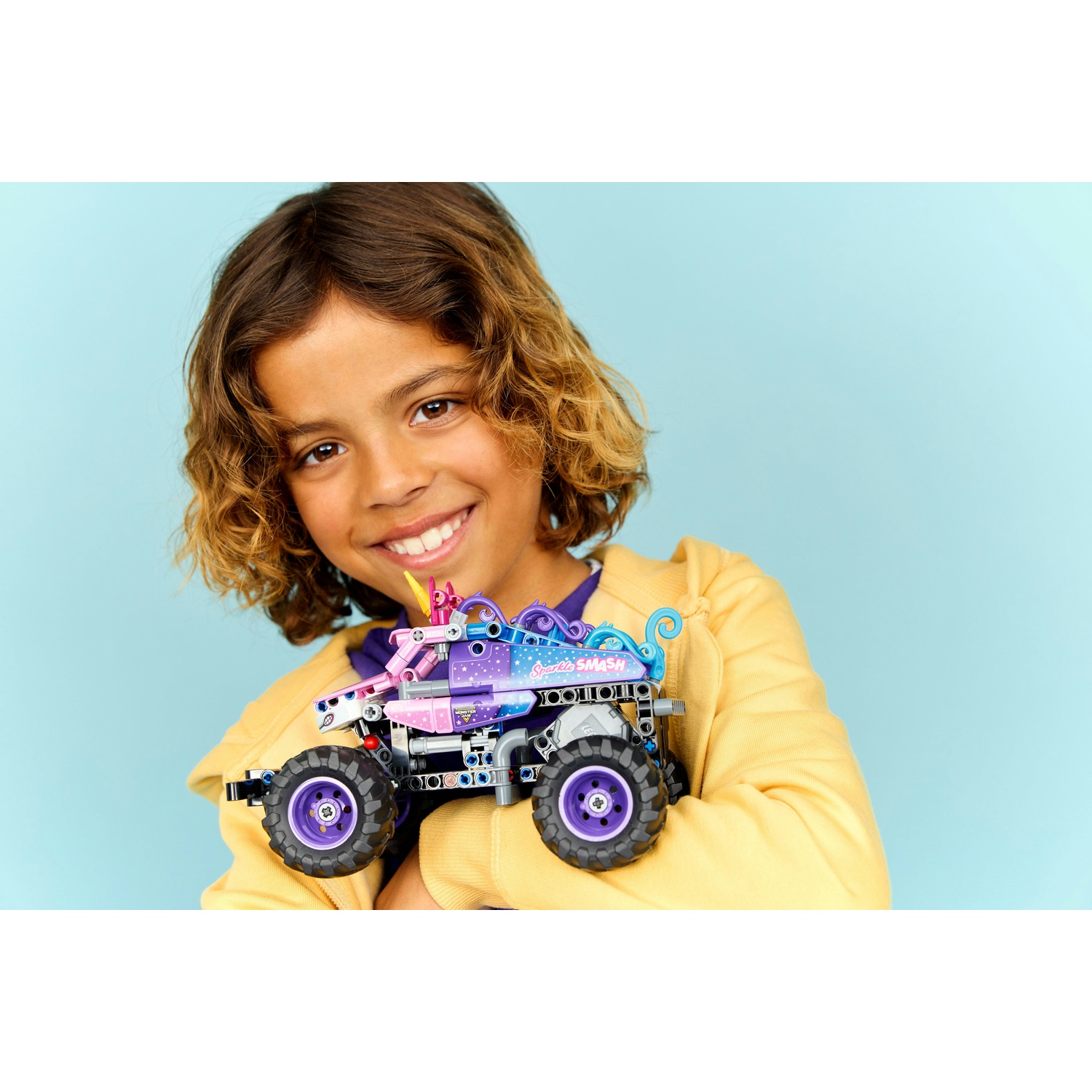 Un bambino sorridente con felpa gialla tiene un monster truck viola su sfondo azzurro.