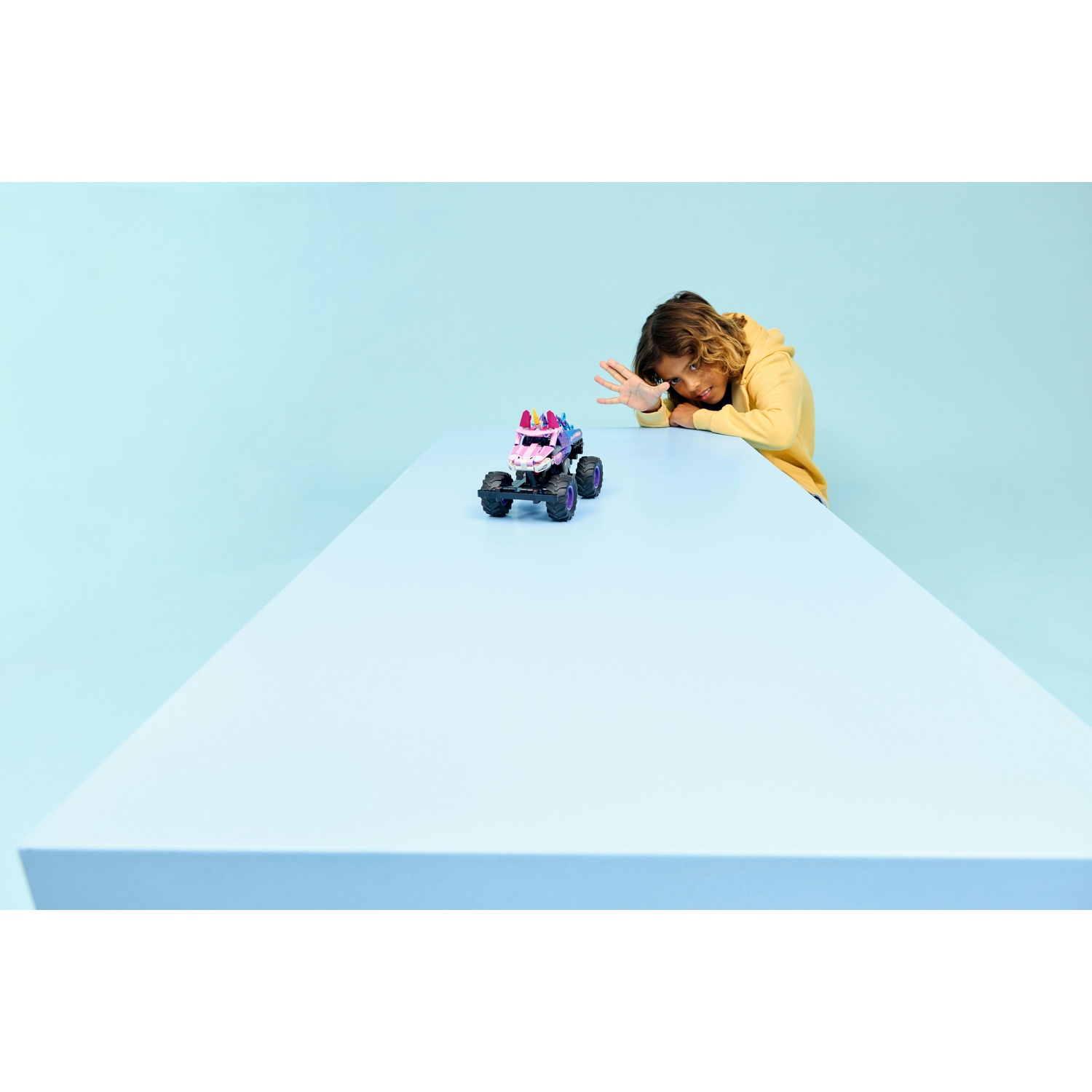Un bambino con felpa gialla guarda oltre un grande tavolo blu con un camion monster rosa.
