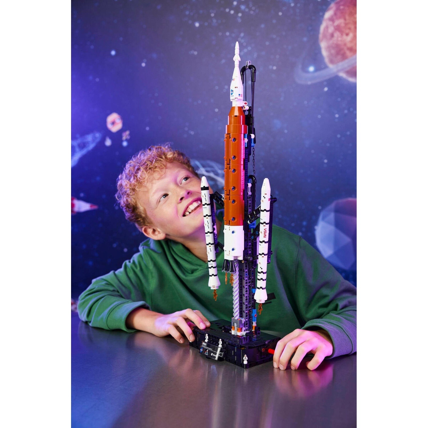 Un bambino biondo con felpa verde guarda orgoglioso un grande modello di razzo Lego davanti a uno sfondo spaziale.