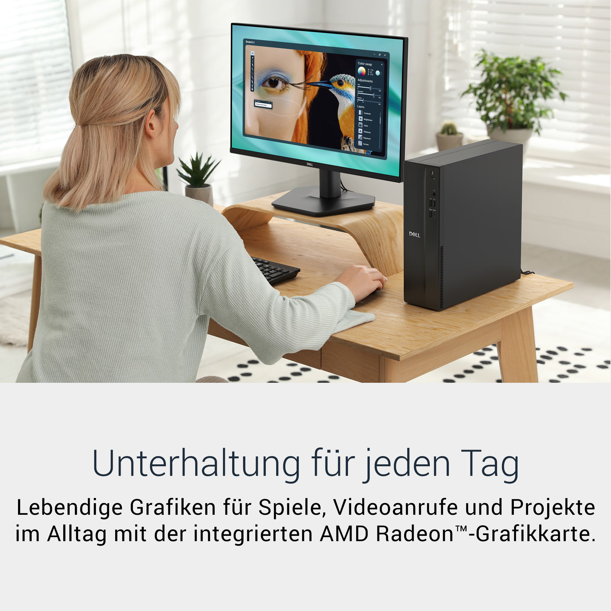 Eine blonde Frau sitzt an einem Holztisch, arbeitet am Computer mit einem Monitor und PC, im Hintergrund Pflanzen.