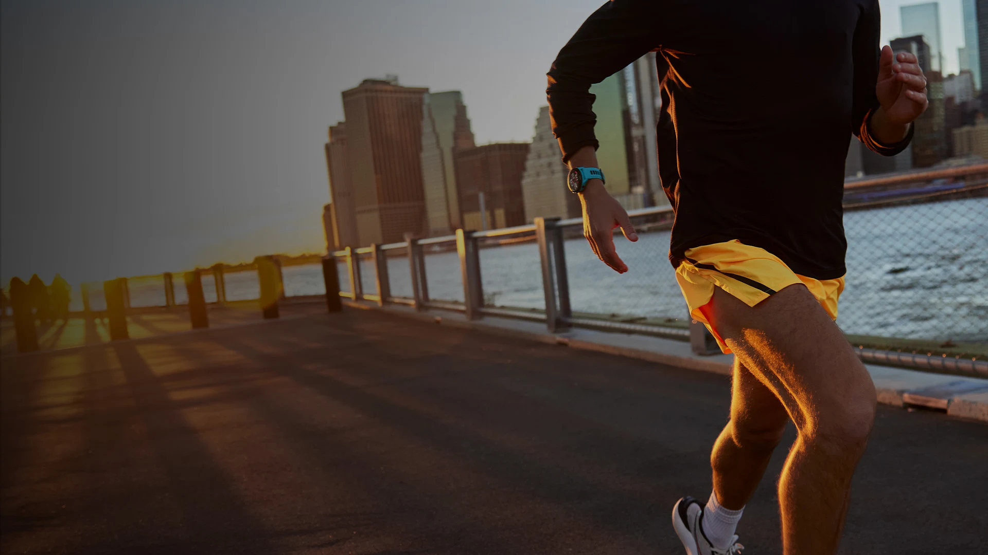 Ein Läufer in schwarzem Shirt und gelben Shorts läuft bei Sonnenuntergang vor einer Skyline am Wasser.