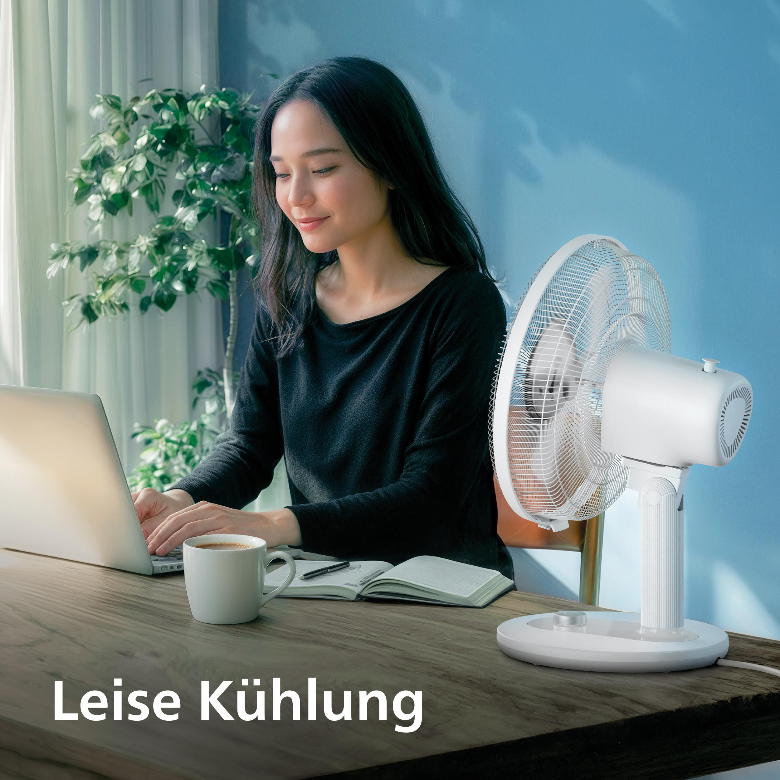 Eine Frau arbeitet am Laptop an einem Holztisch, neben ihr steht ein weißer Ventilator.